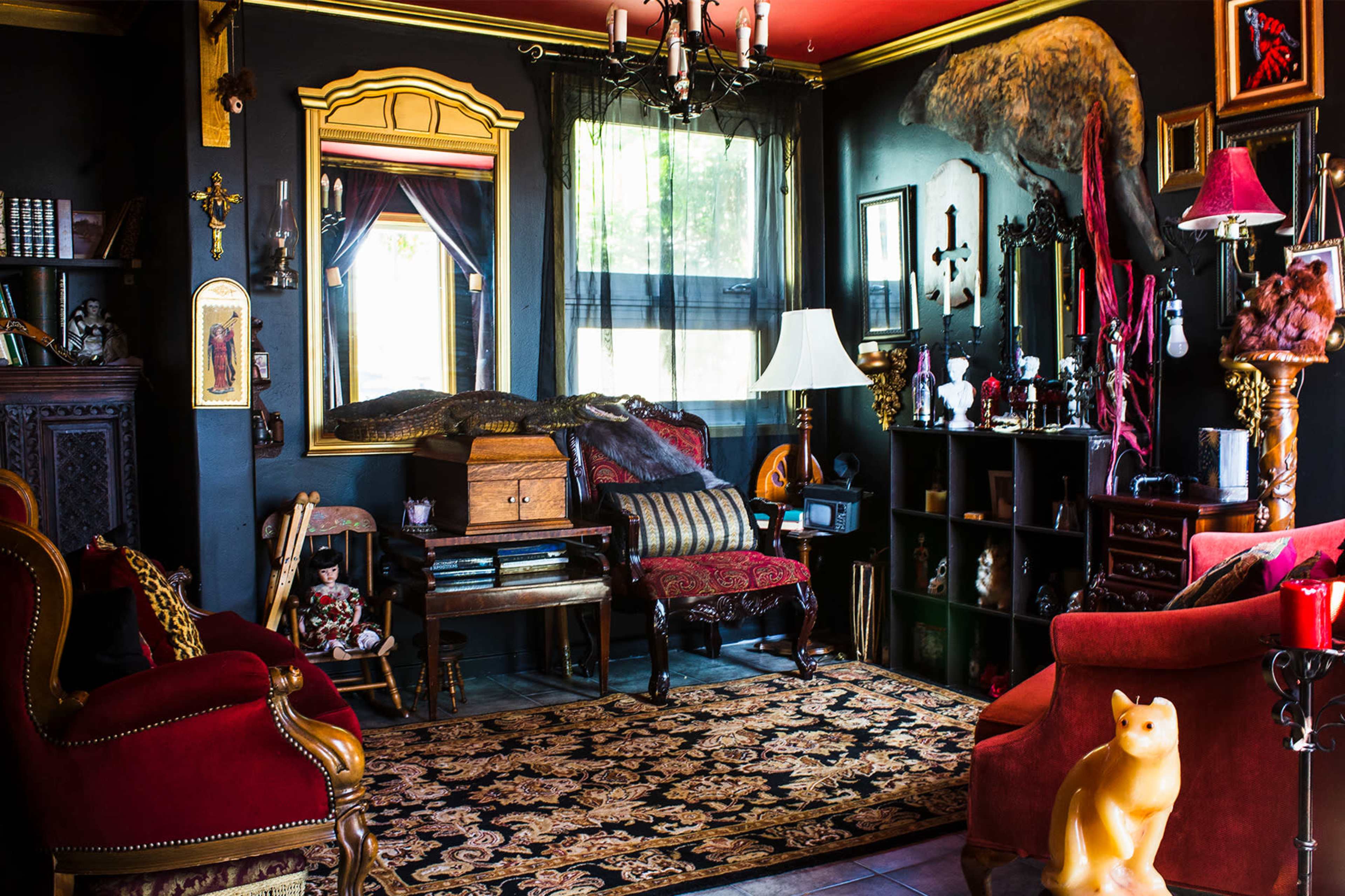 The room features a mix of antique furniture, dark walls adorned with diverse decor, a piano, and a large carpet under a chandelier.