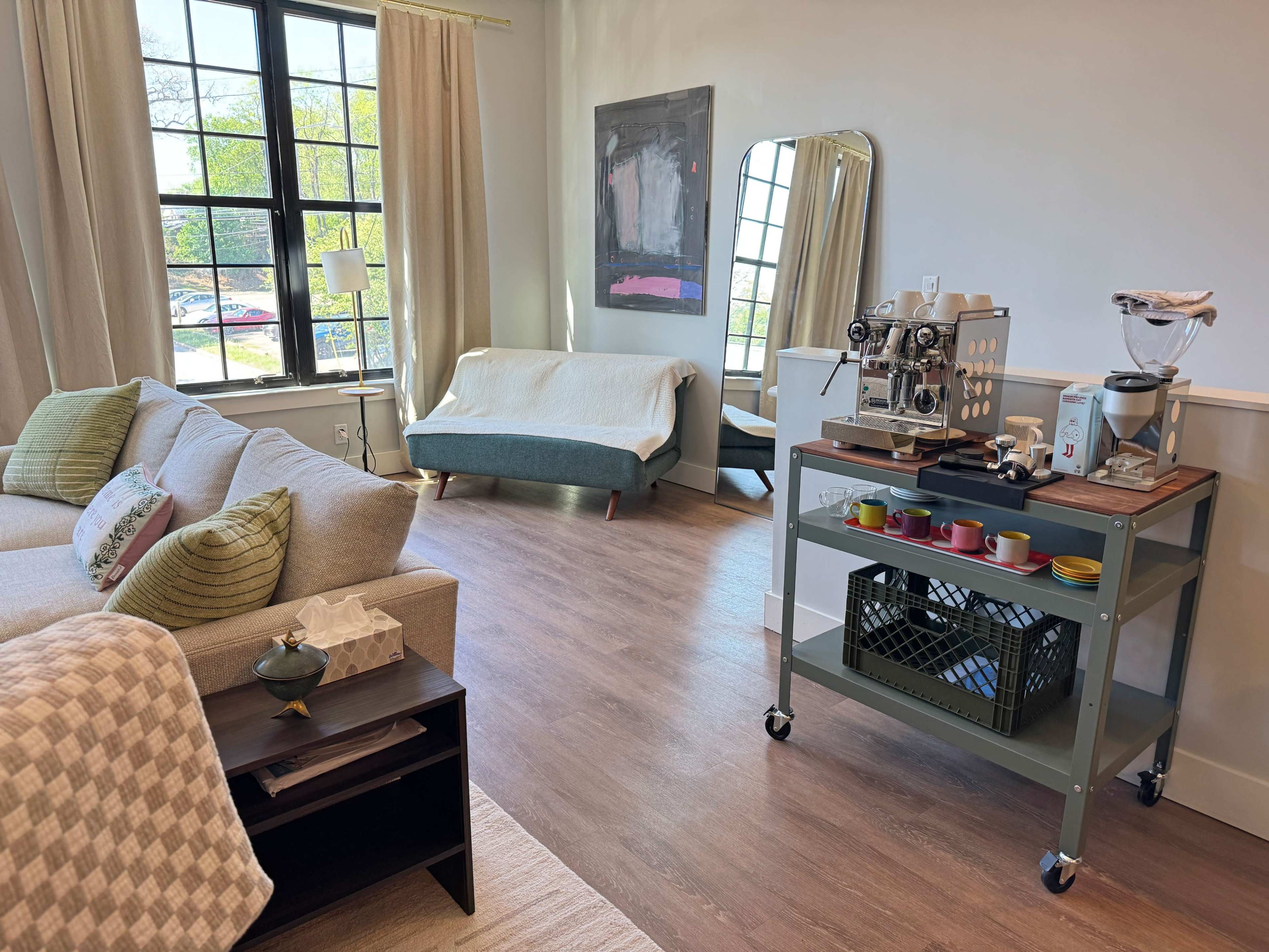 The image shows a cozy, modern living space featuring a sofa, a large mirror, and a coffee cart with colorful cups.