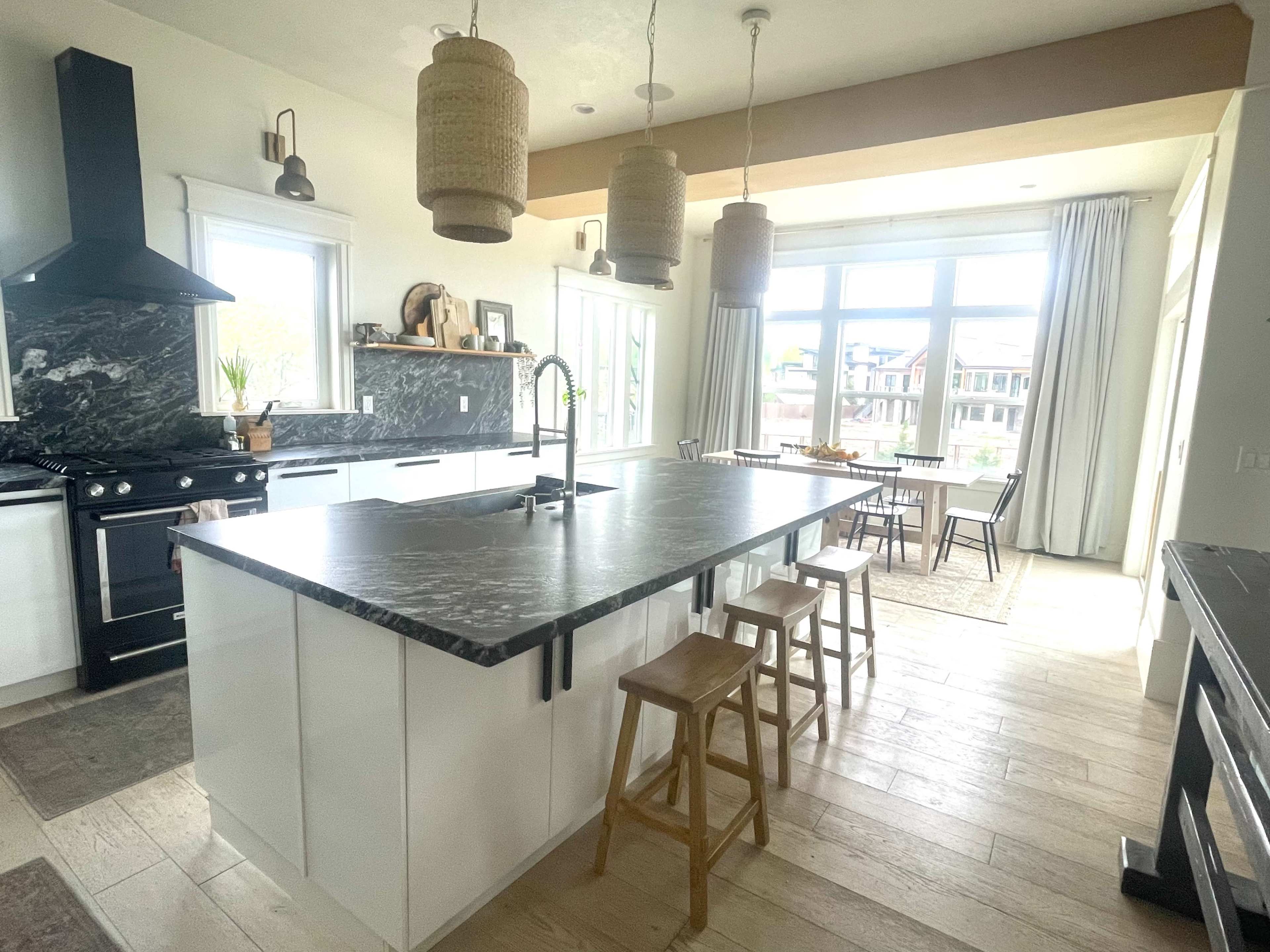 A modern kitchen features a large island with a dark countertop, surrounded by wooden stools and illuminated by pendant lights, with a dining area and large windows in the background.