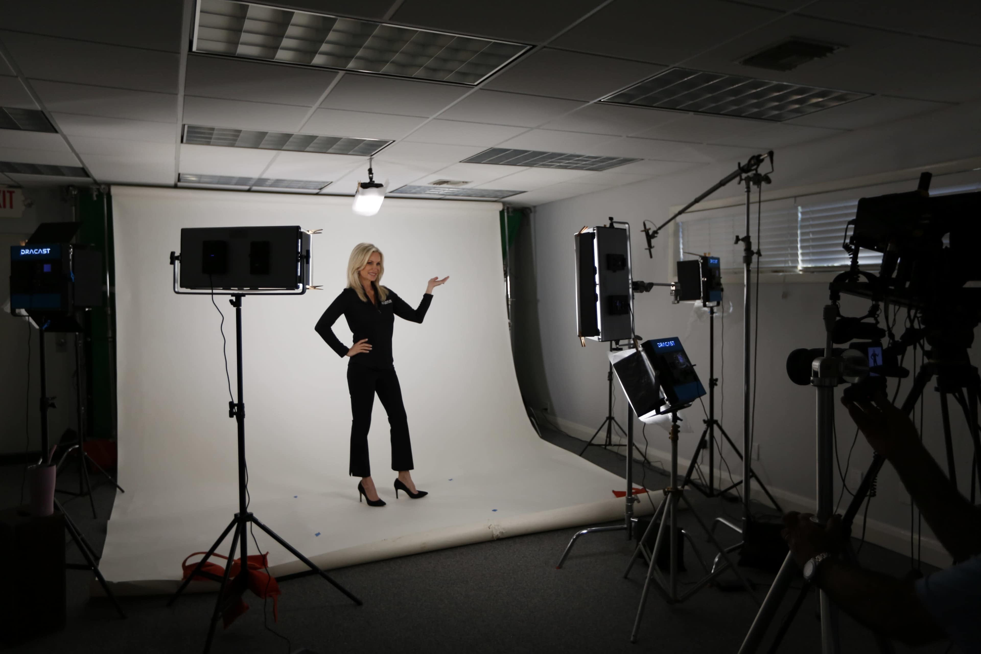 A woman in a black outfit poses with one hand raised in a photography studio surrounded by lighting equipment and a backdrop.