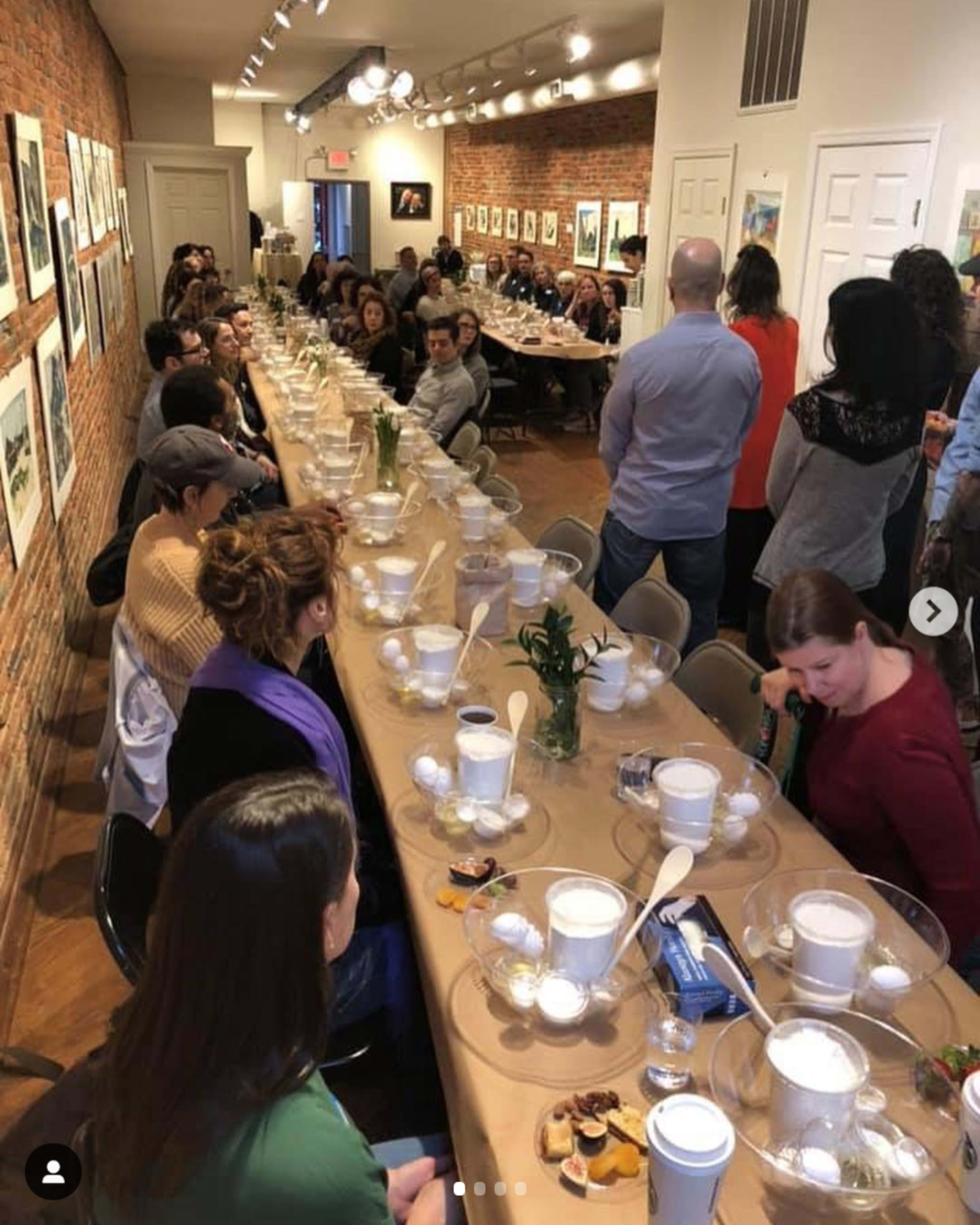 A long table is set for a gathering, with attendees seated on either side, surrounded by artwork hung on the brick walls.
