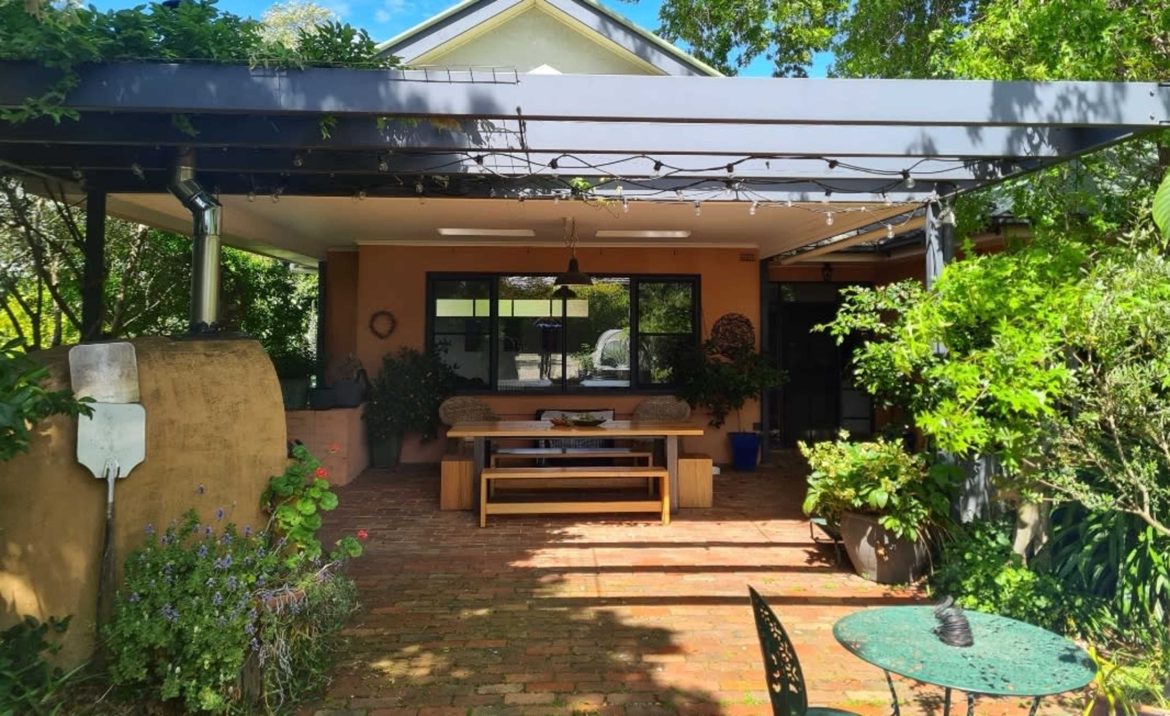 A covered outdoor patio features a wooden table and benches surrounded by greenery and decorative plants.