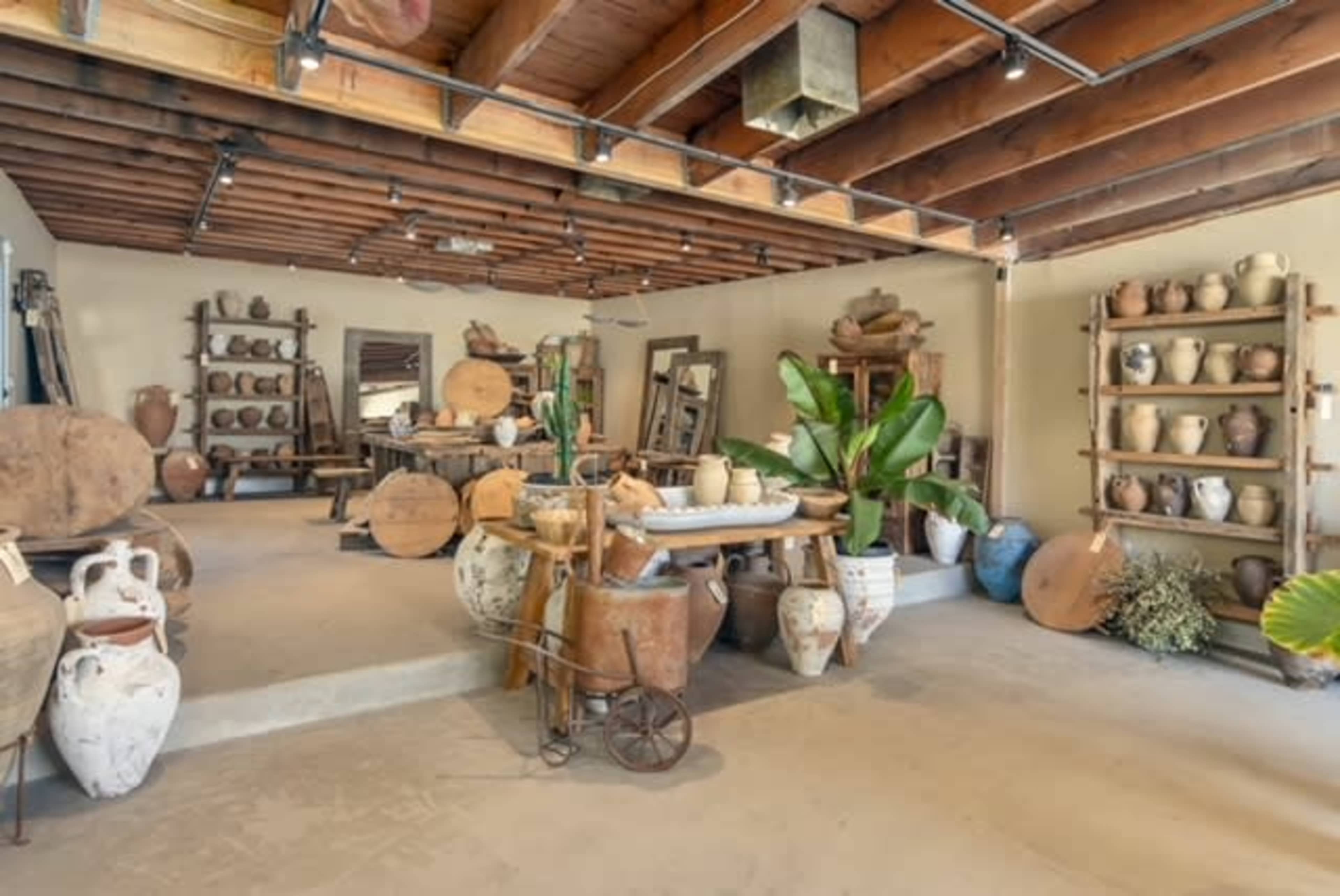 The image shows an interior space filled with various pottery pieces, wooden furniture, and decorative plants against a backdrop of exposed wooden beams.
