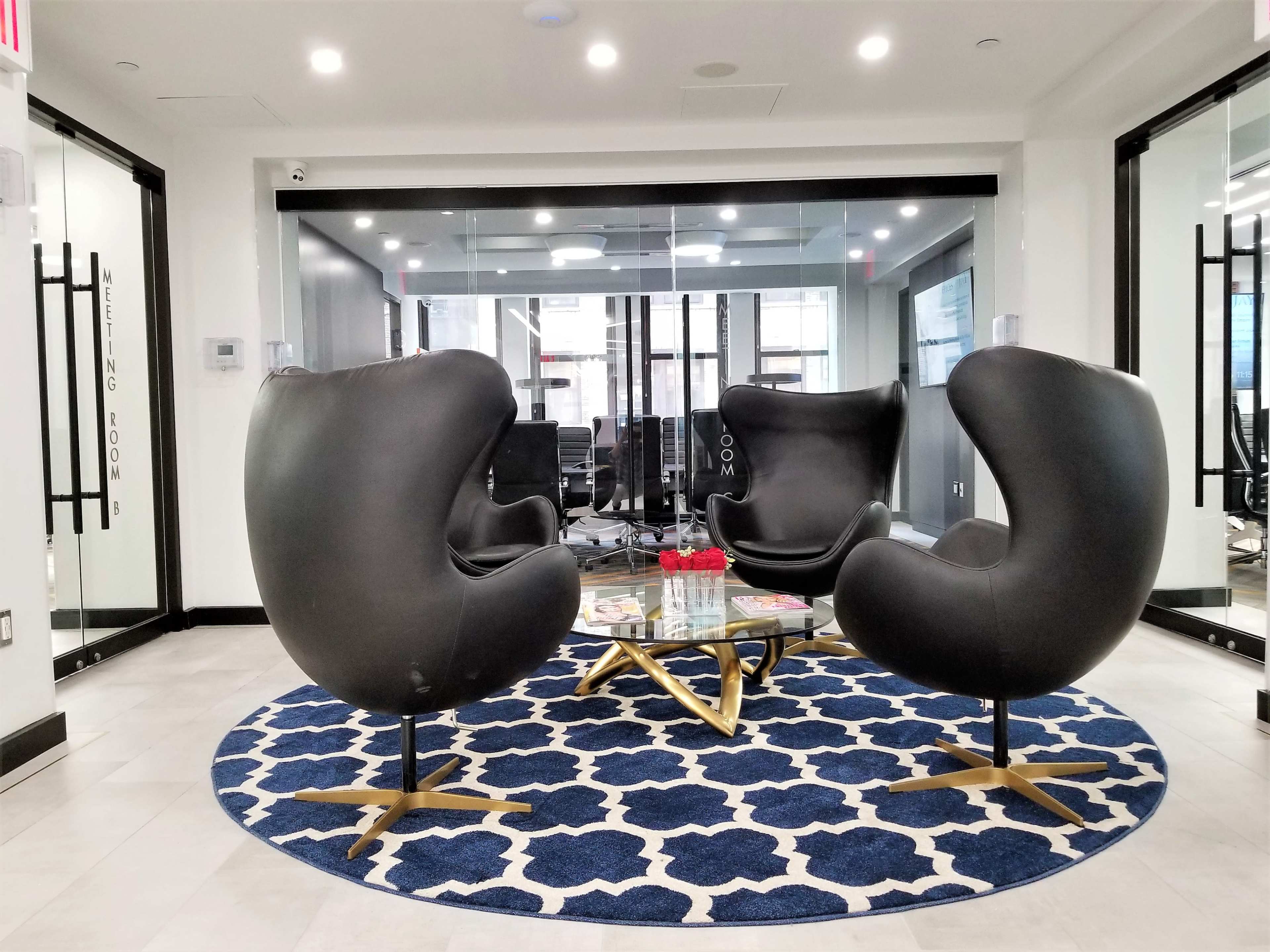 A modern office space with two black chairs positioned around a glass table on a patterned rug, surrounded by glass walls and meeting rooms.