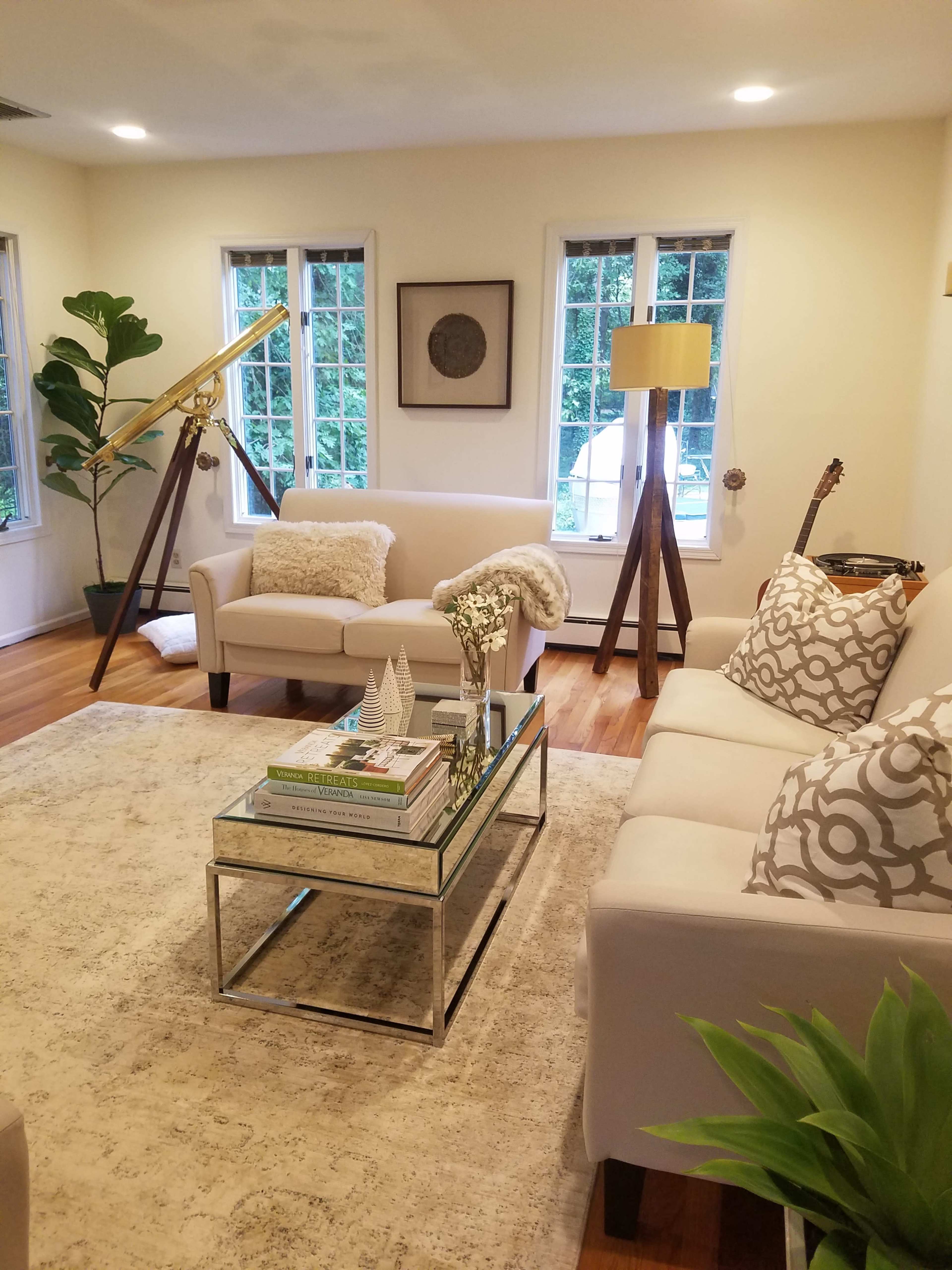 The image depicts a cozy living room featuring a light-colored sofa, a glass coffee table with decorative items, a guitar, and large windows allowing natural light to enter.
