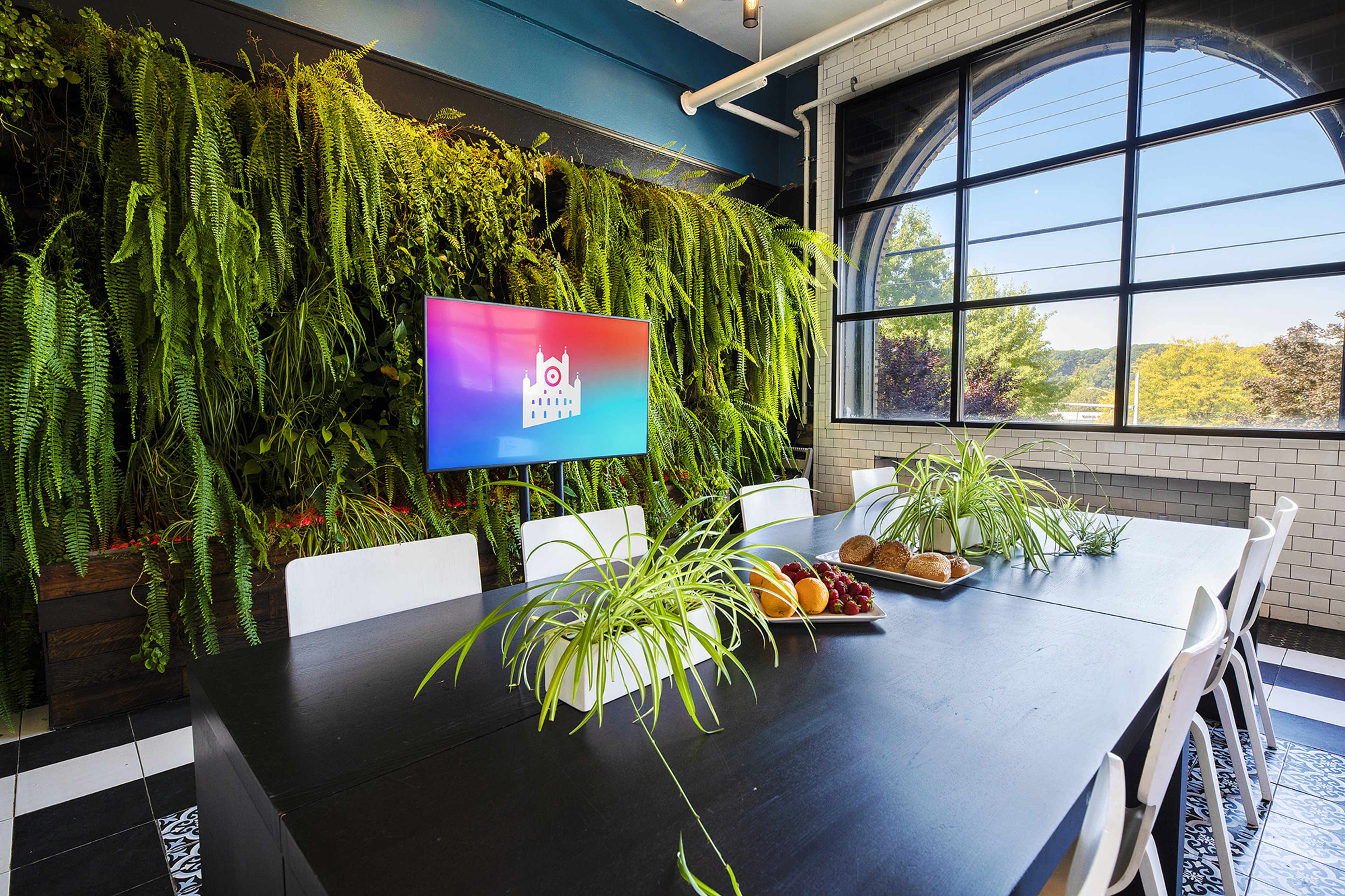 A large meeting room features a wall of greenery, a digital screen, and a dining table set with fruits and baked goods.
