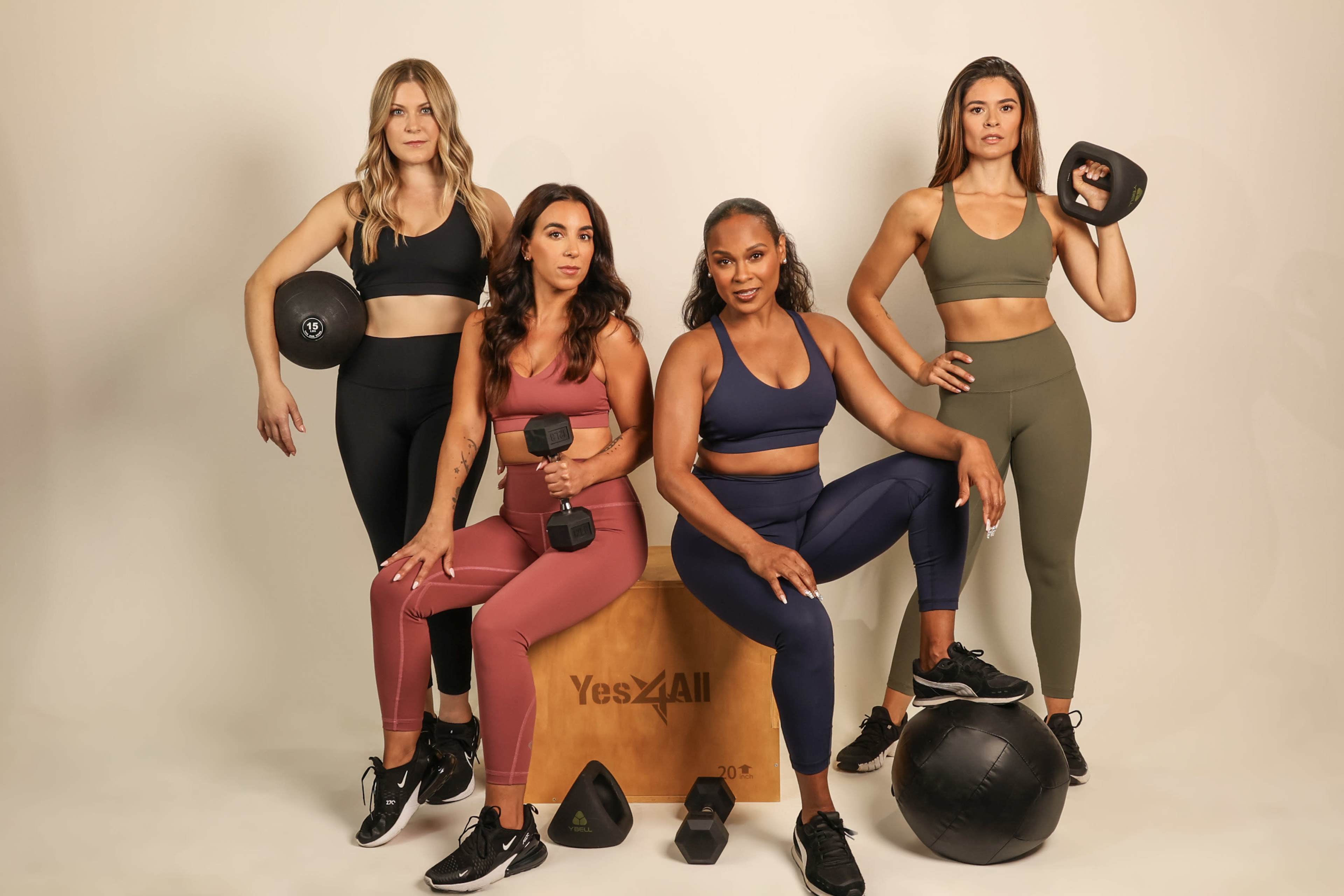 Four women in activewear pose with various fitness equipment, including dumbbells and a kettlebell, in front of a wooden box.
