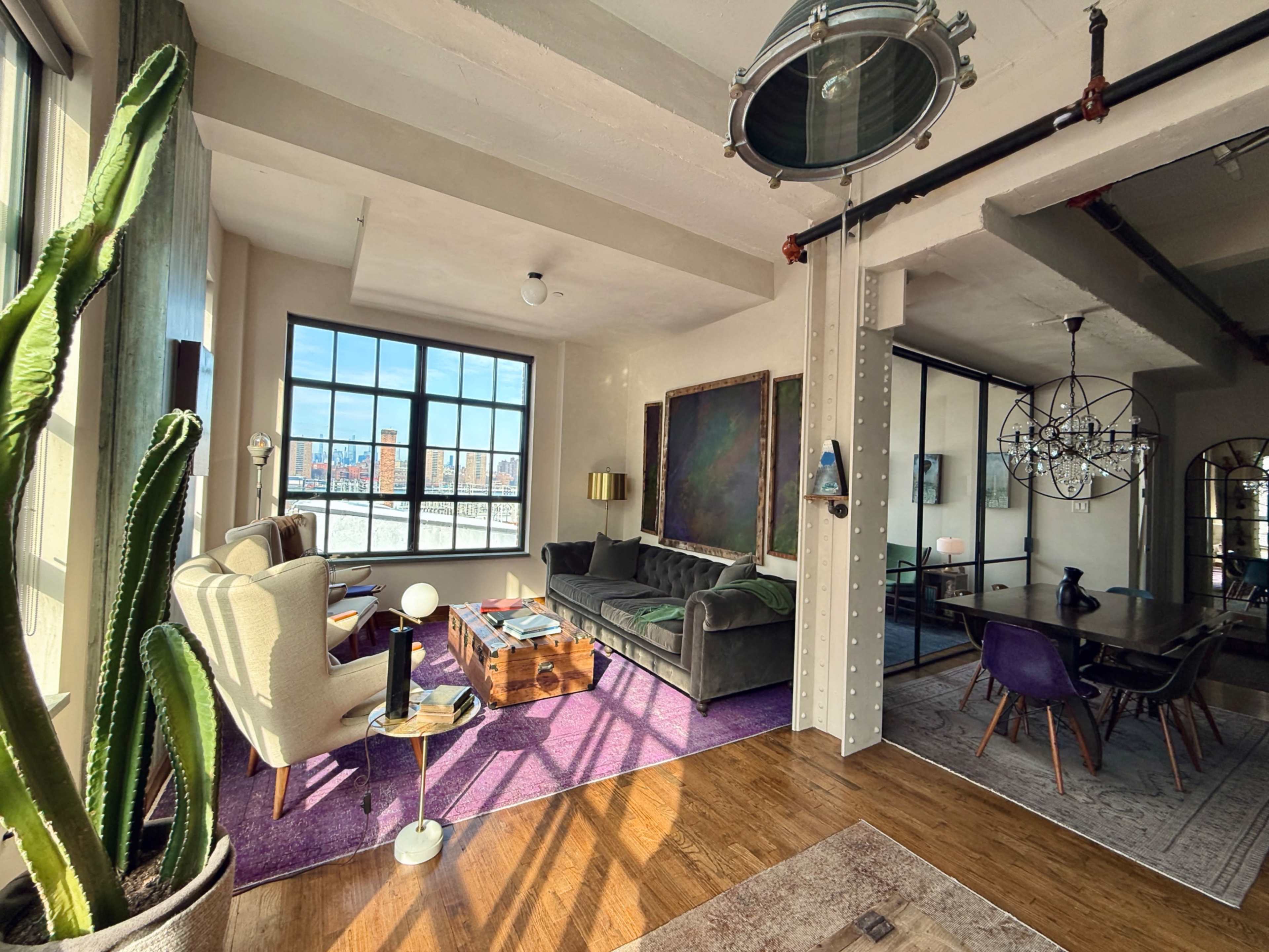 The image shows a spacious, modern living area with large windows, featuring a mix of furniture including a couch, armchairs, and a coffee table on a purple rug.