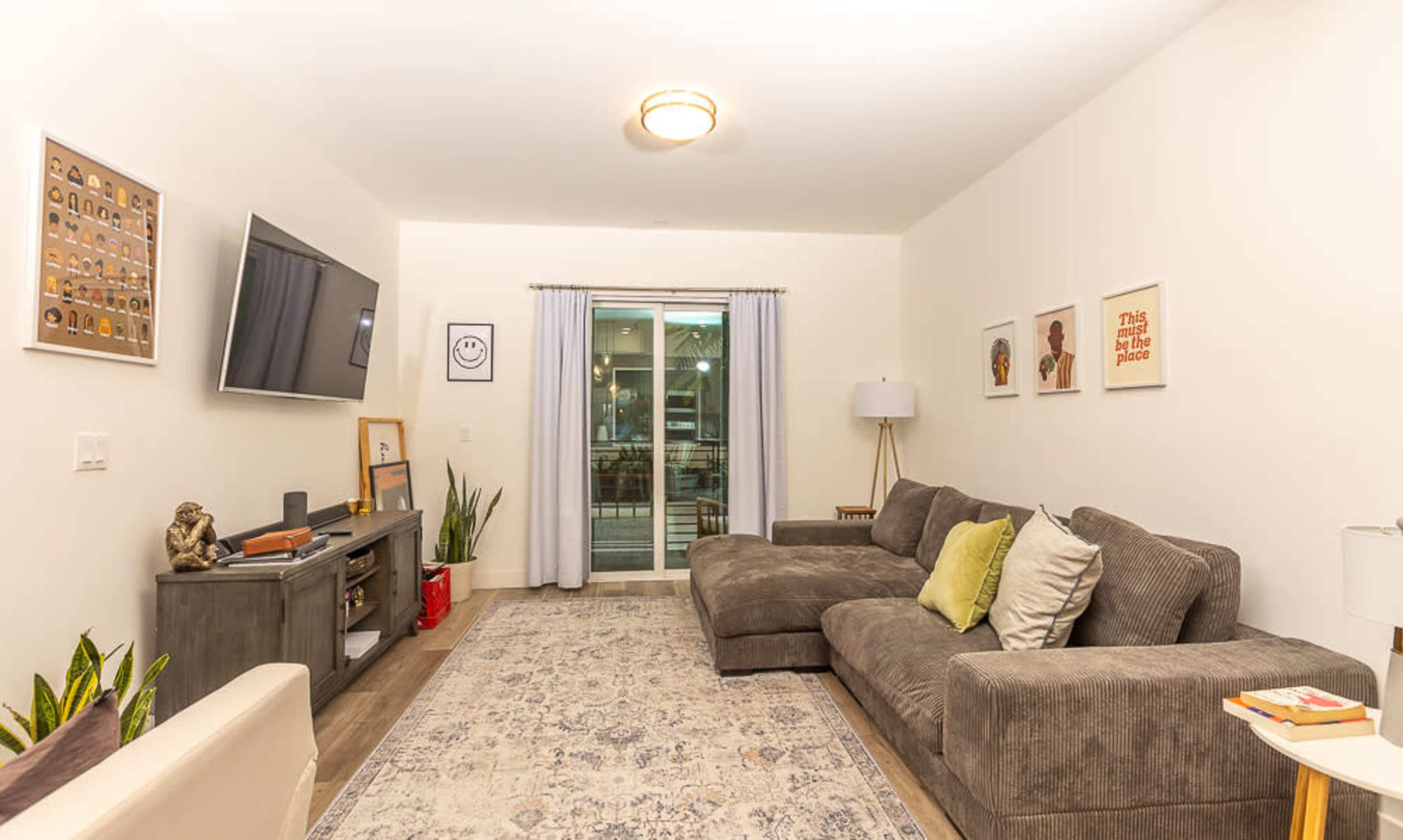 A modern living room with a large sectional sofa, a television mounted on the wall, a sliding glass door leading outside, and various decorative elements including plants and framed art.