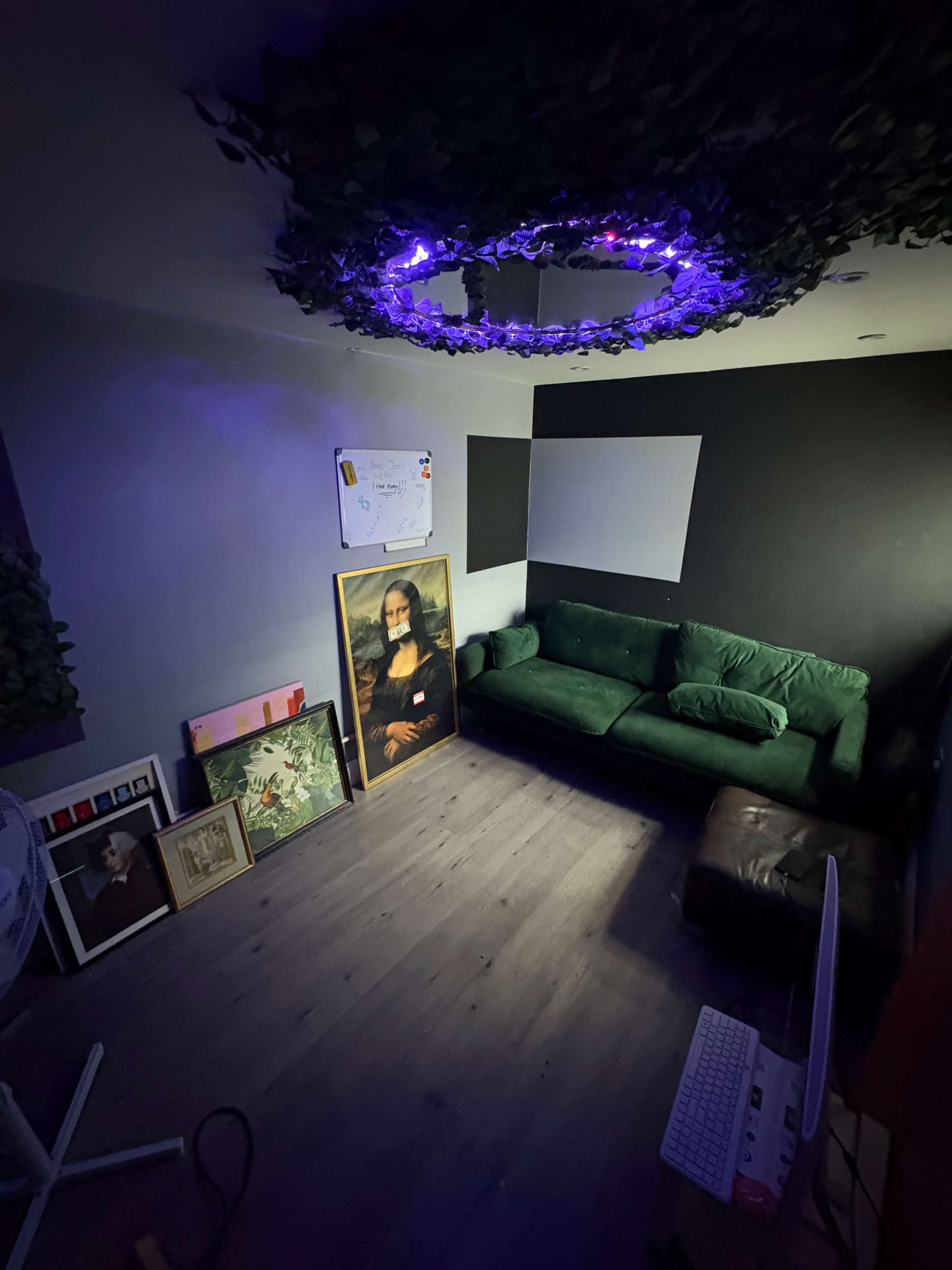 The image shows a dimly lit room with a green sofa, a large portrait on the floor, framed artworks on the wall, and a ceiling decorated with leaves and purple lights.