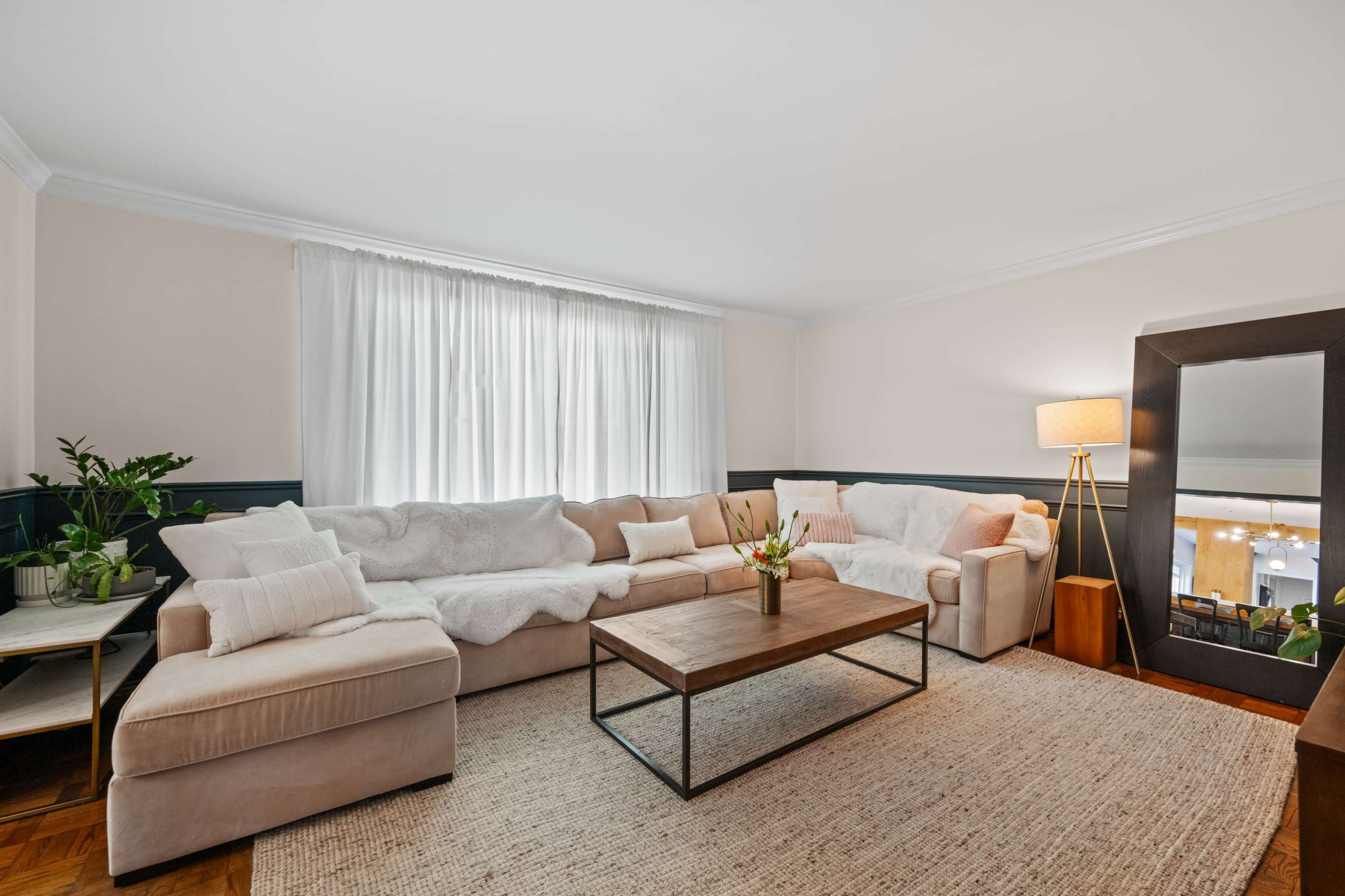 A cozy living room with a light-colored sectional sofa, a wooden coffee table, and a large mirror beside a floor lamp, all set against a backdrop of large windows with sheer curtains.