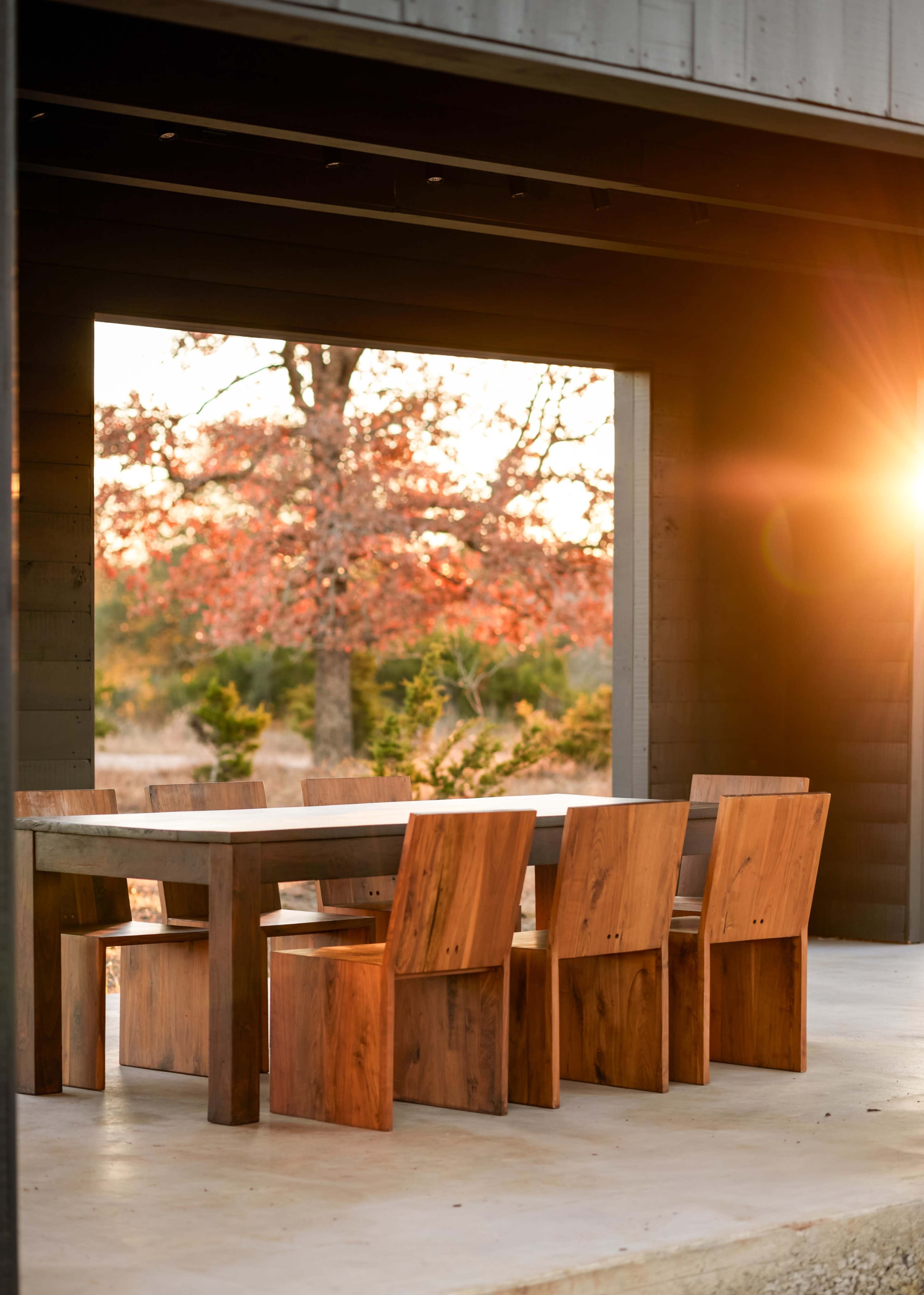 A wooden dining table is set with six matching chairs in a modern outdoor space, framed by a large opening that views a sunset over autumn foliage.