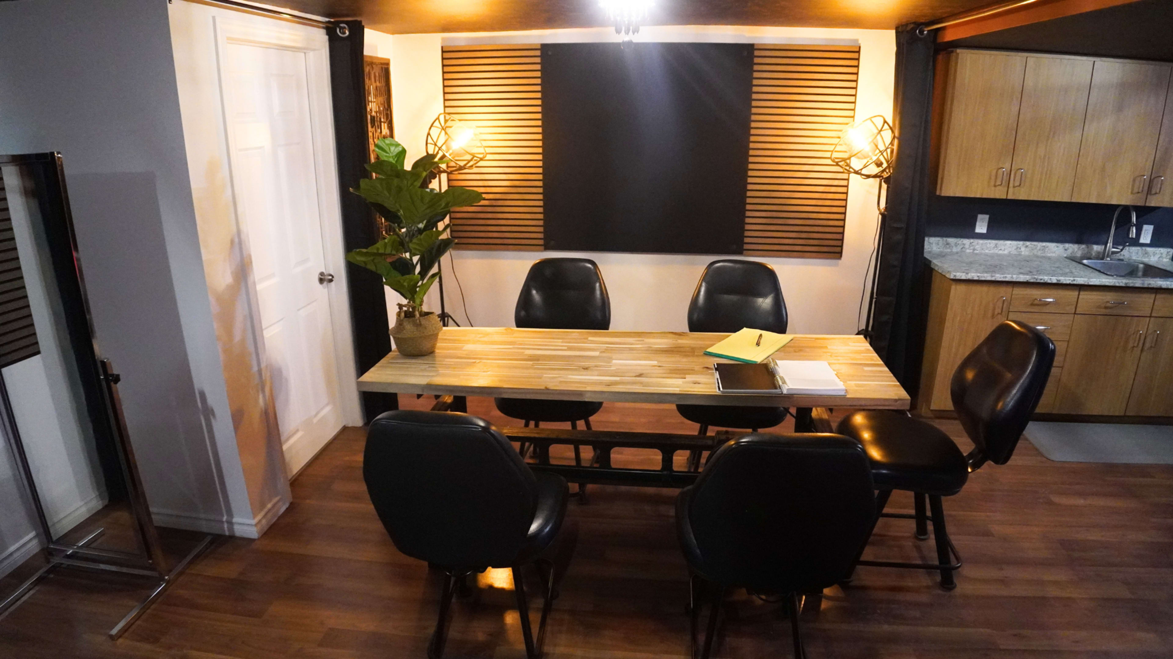 A conference room features a wooden table surrounded by black chairs, illuminated by overhead lighting, with a kitchenette and decorative plant in the background.