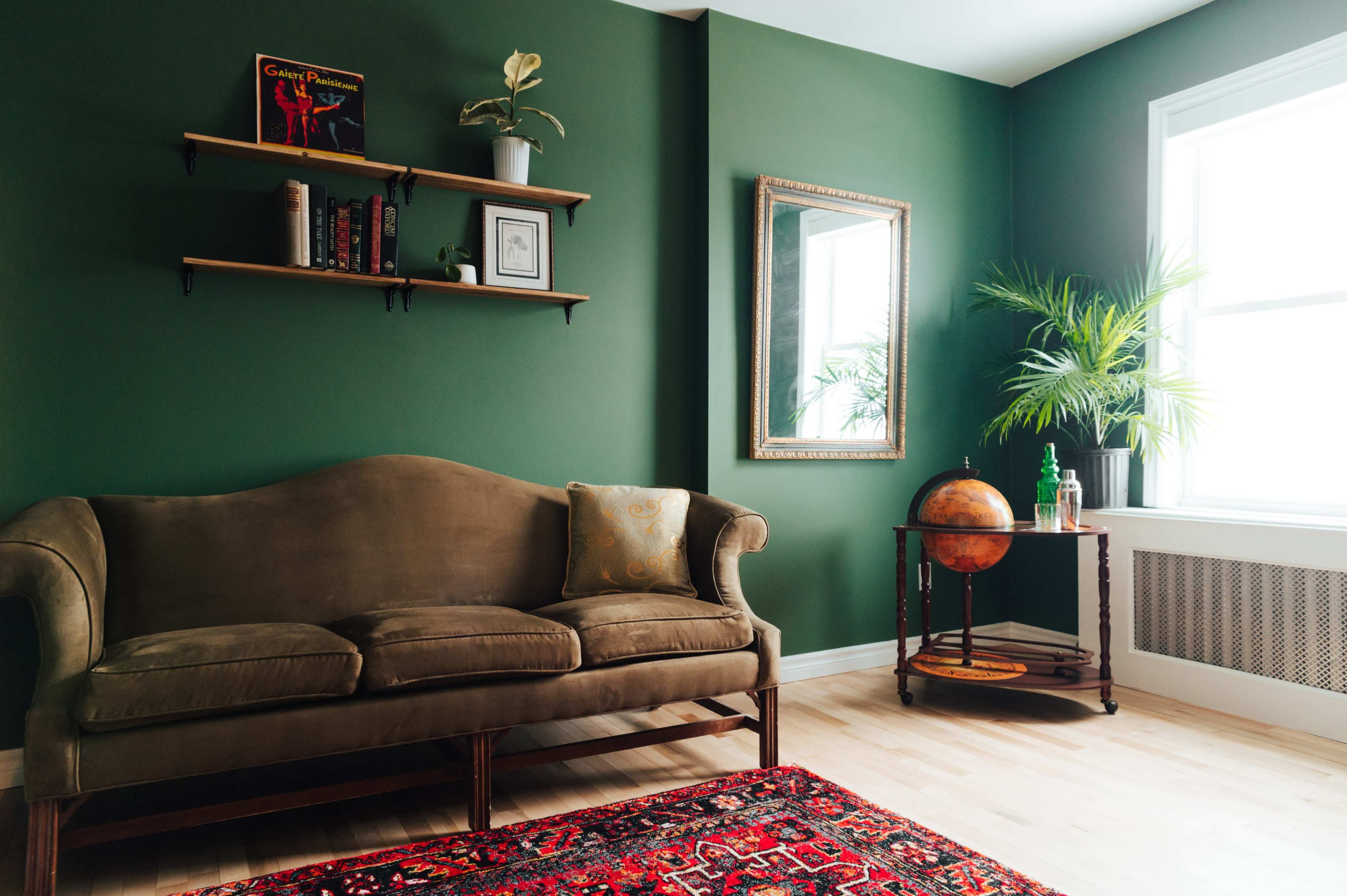 A living room with a green wall, a brown couch, a wooden shelf with books and a plant, a mirror, and a globe on a decorative stand.