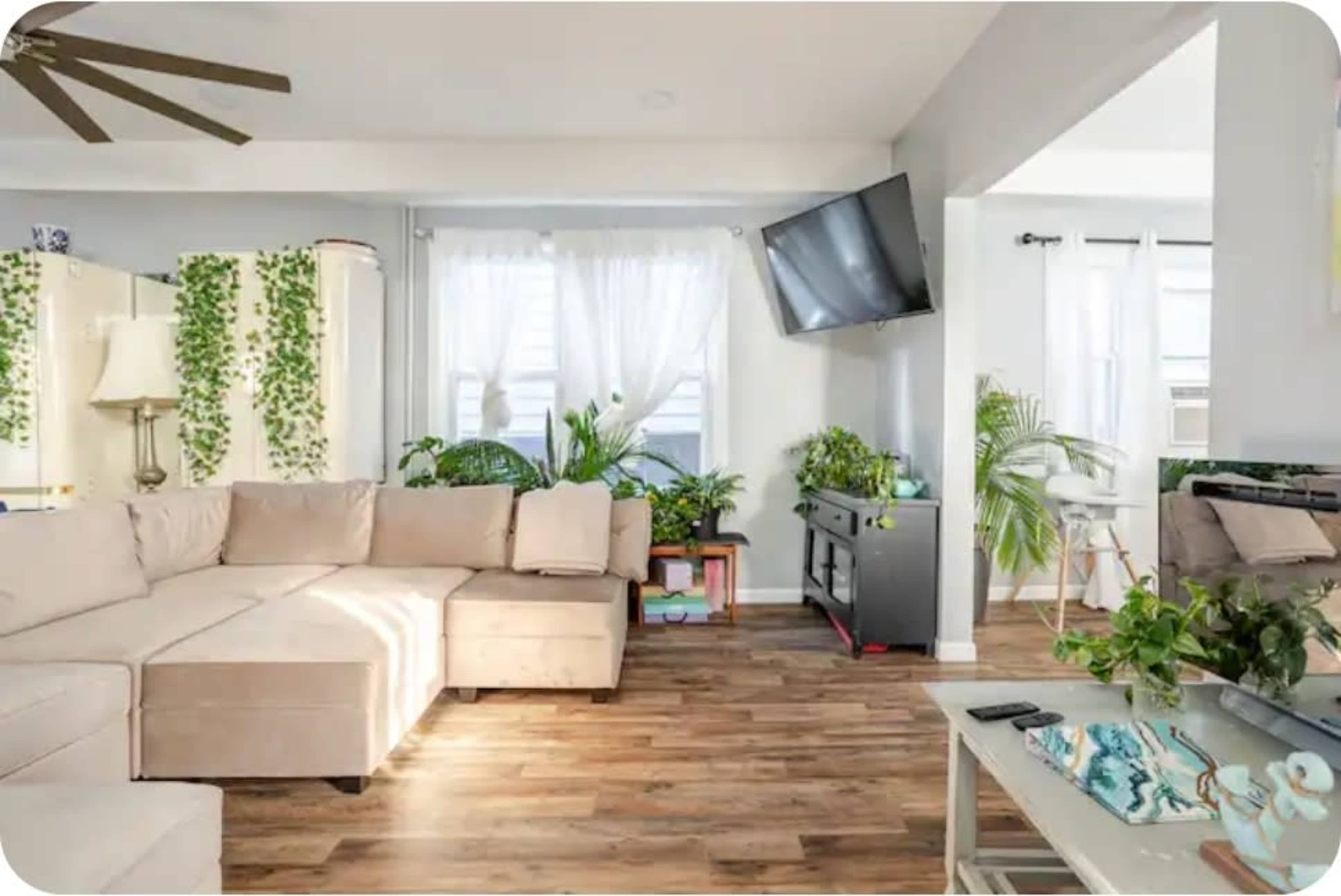 The image depicts a living room with a sectional sofa, large windows letting in natural light, and various indoor plants arranged throughout the space.