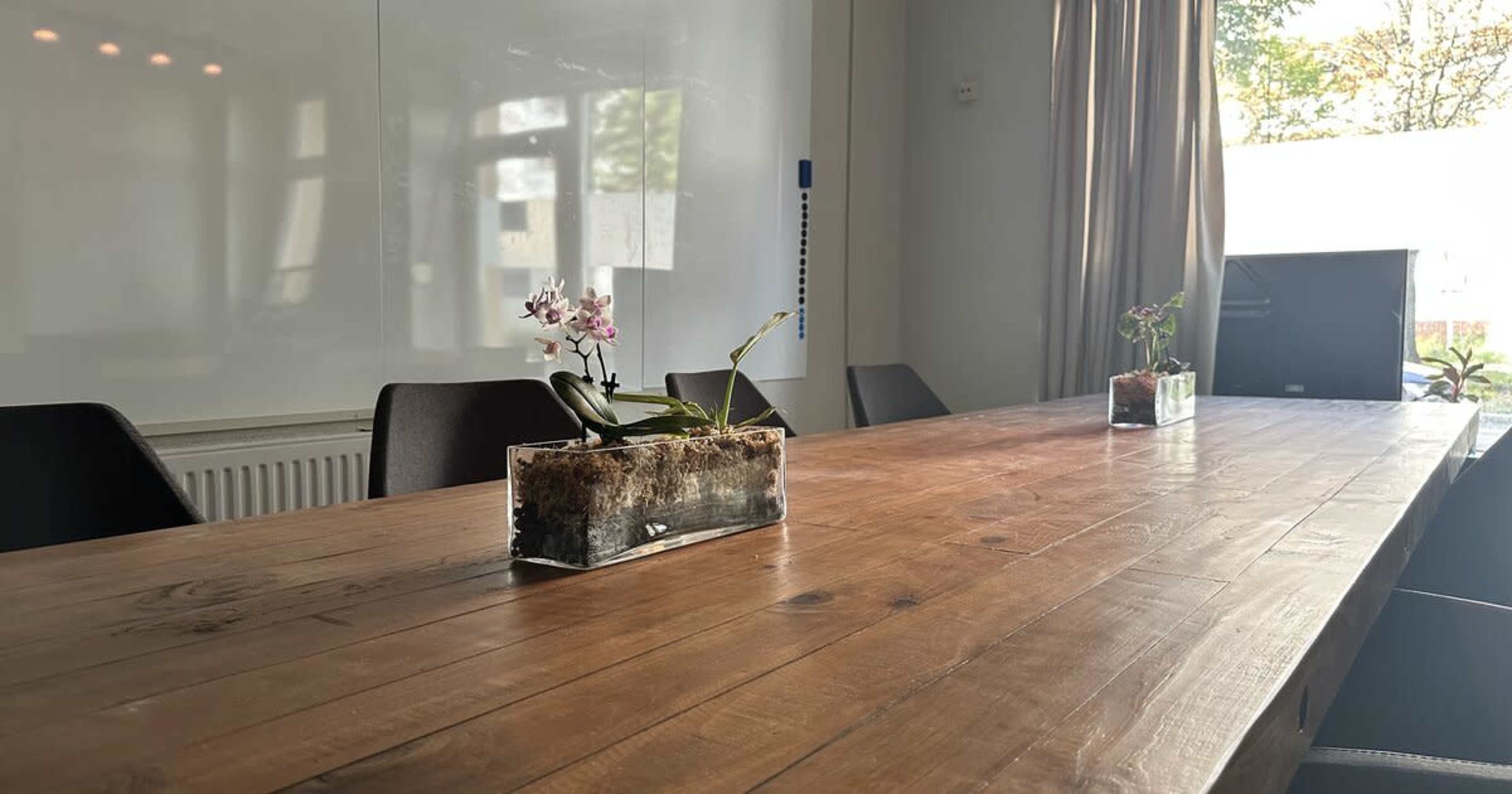 A long wooden table is set in a well-lit conference room with two glass vases containing plants.
