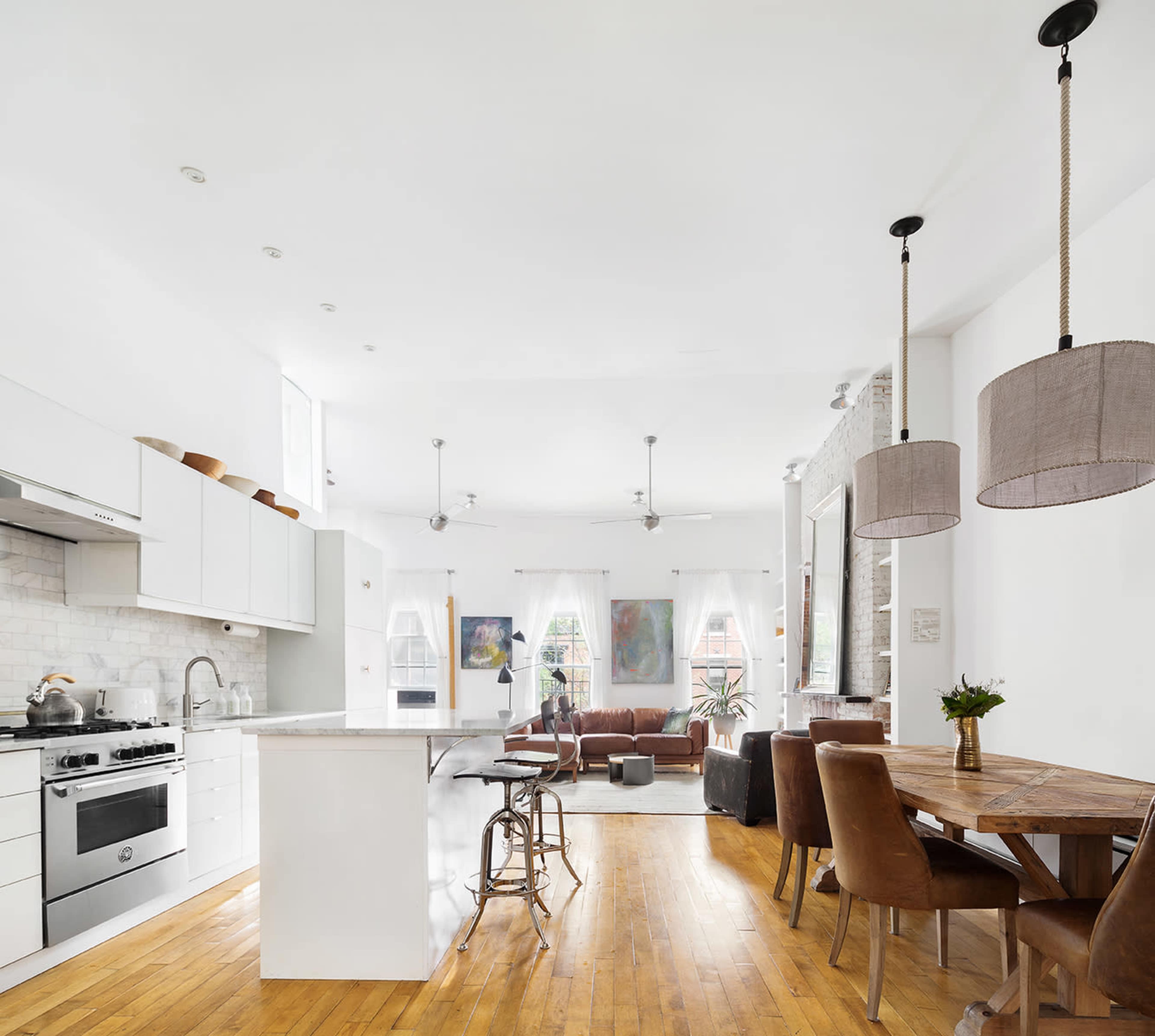 A spacious kitchen and living area featuring modern appliances, wooden floors, a dining table, and large windows with natural light.