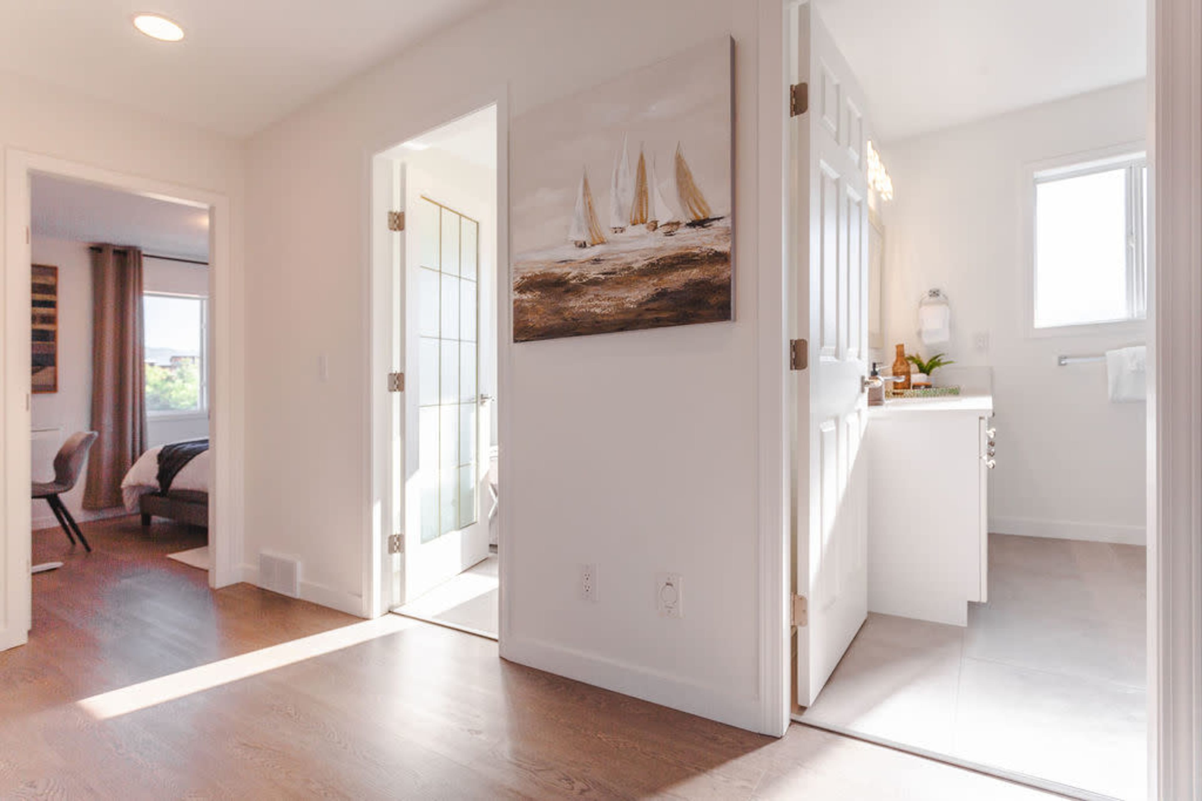 A well-lit hallway connects a bathroom and a bedroom, featuring a painting of sailboats on the wall.