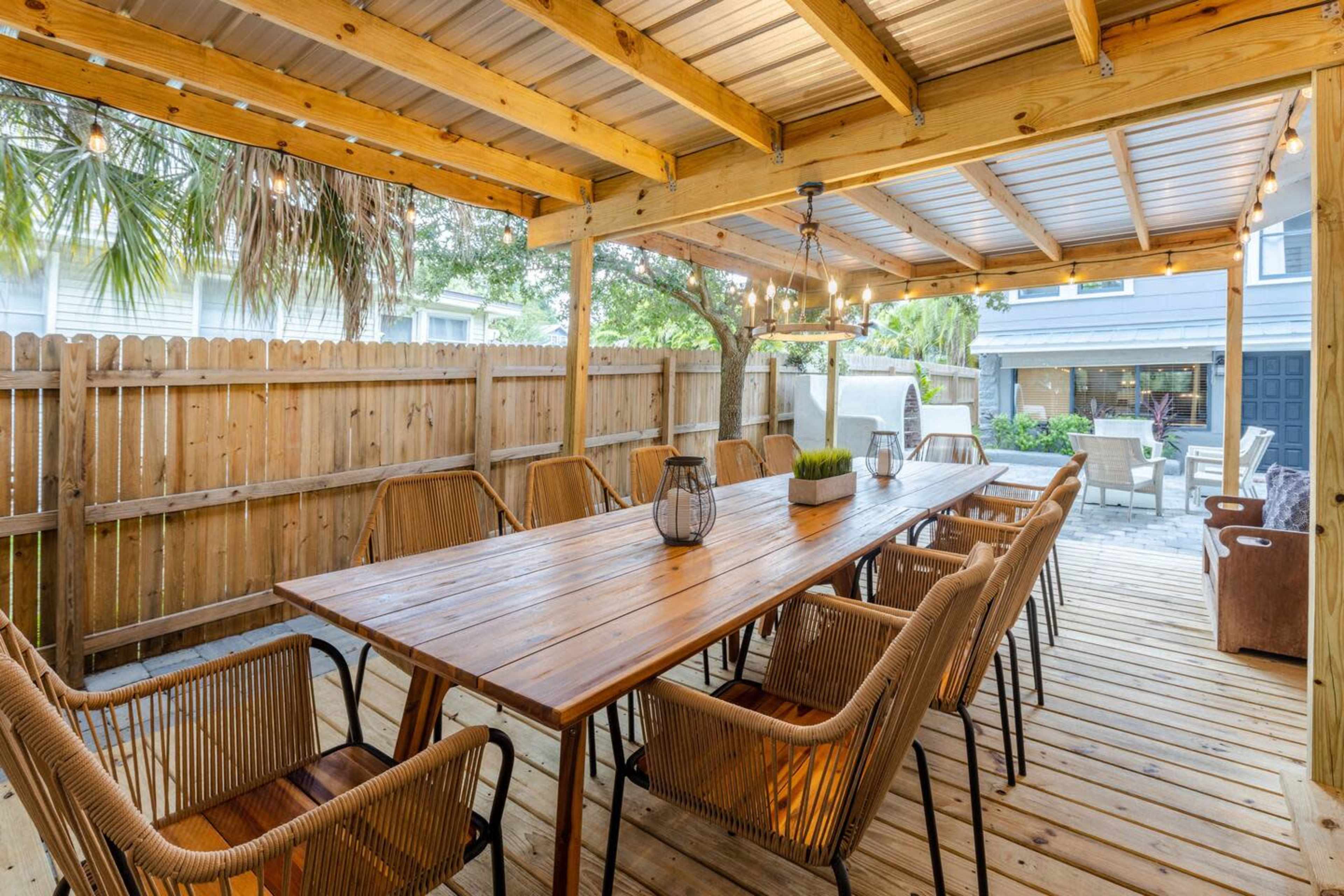 A wooden patio with a long table surrounded by chairs, under a covered structure with string lights.