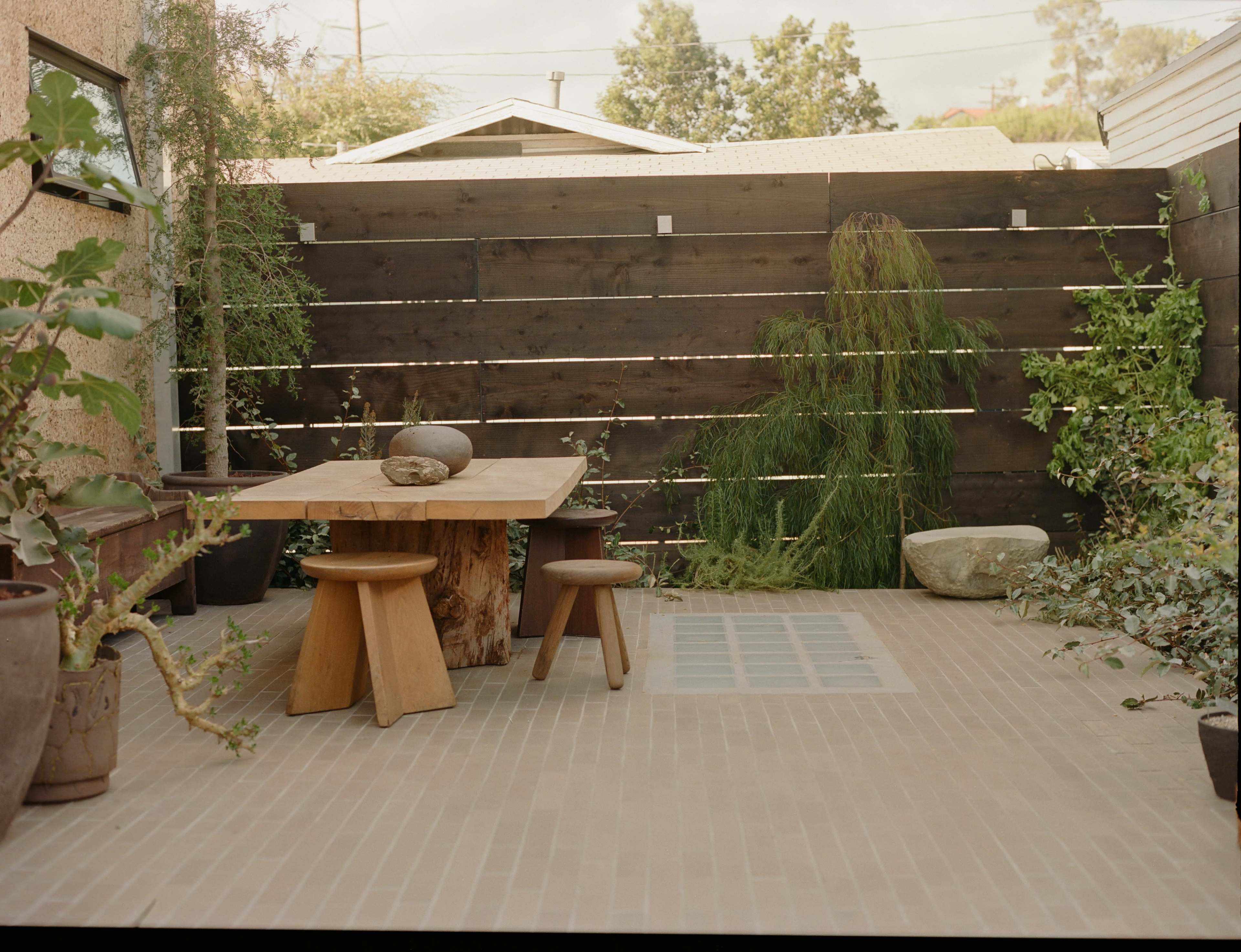 A small outdoor patio featuring a wooden table with three stools, surrounded by potted plants and a wooden fence.