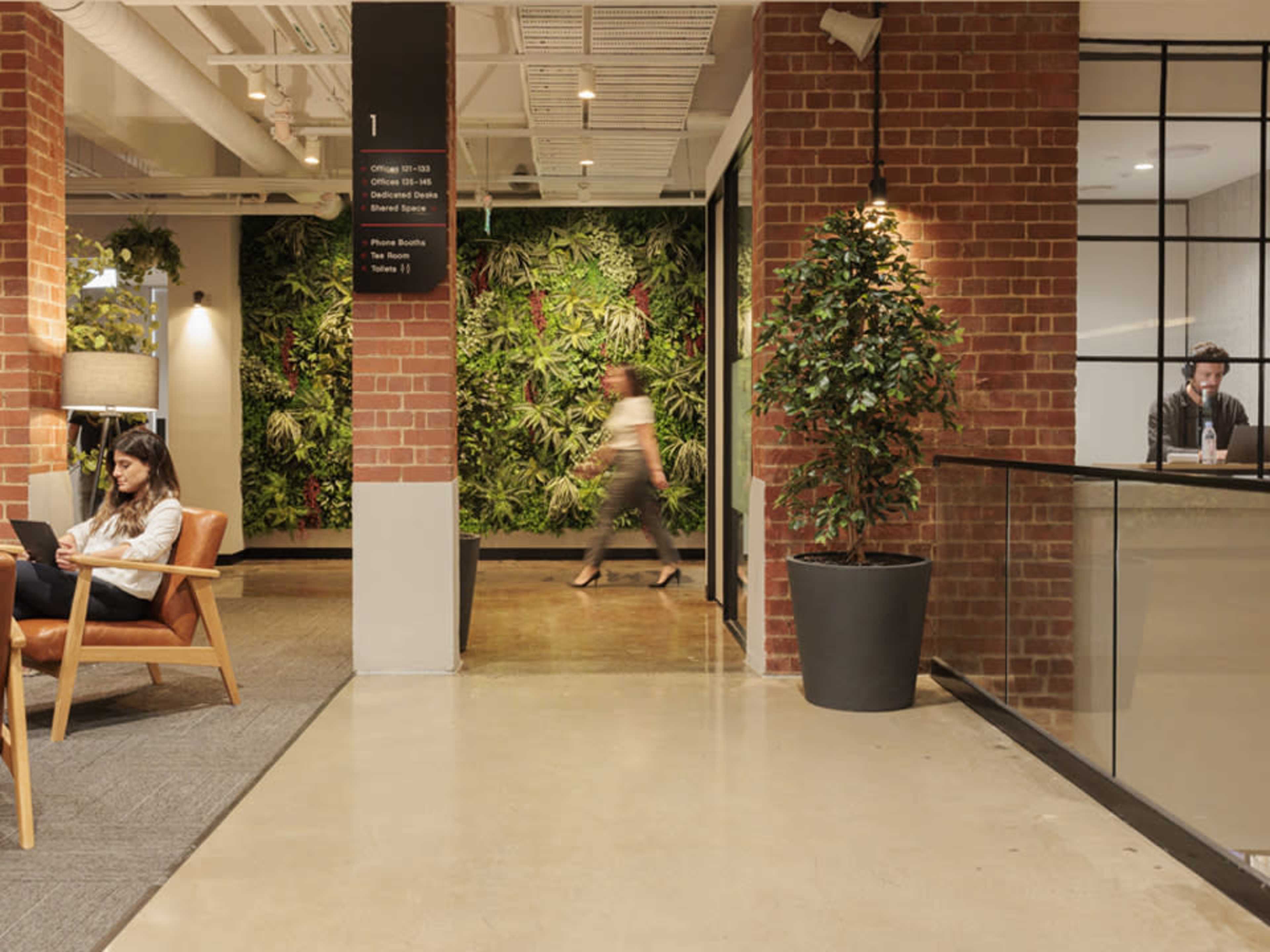 A modern office space featuring a seating area, a green wall with plants, and individuals working at desks.