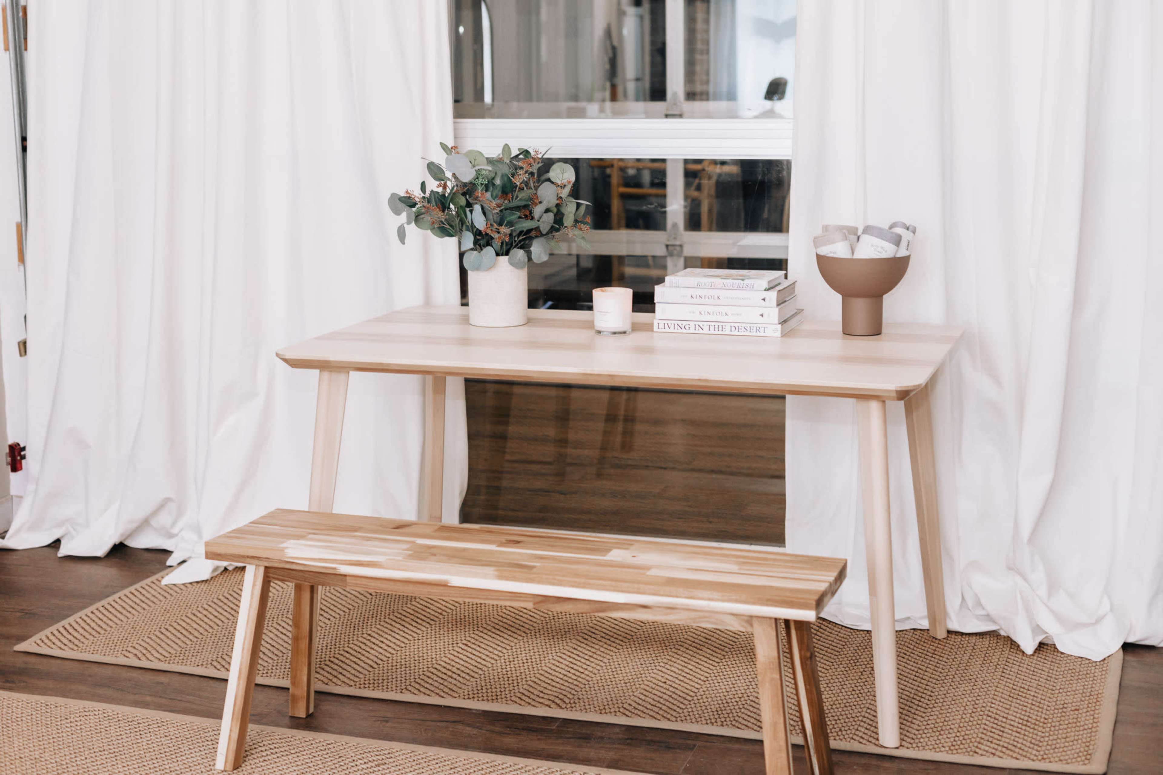 A modern wooden table with a bench sits on a woven rug, featuring a vase of greenery and a stack of books beside a candle.