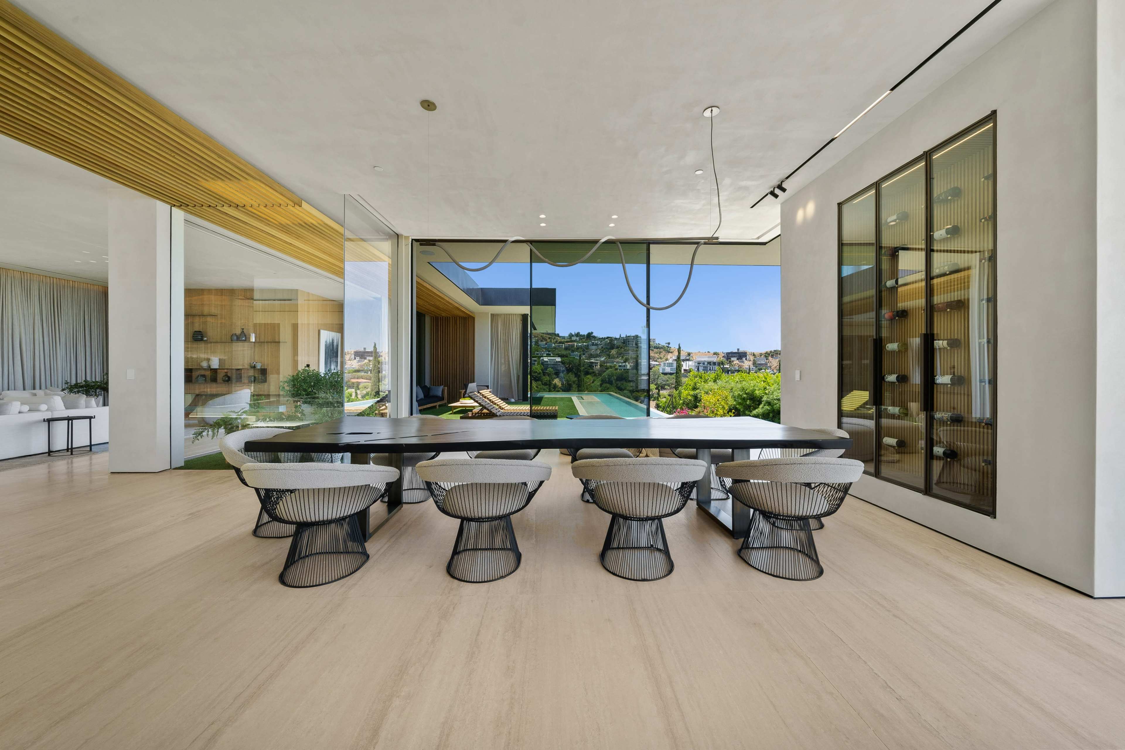 A modern dining area features a long table surrounded by six wireframe chairs, with large glass windows providing a view of a hillside landscape.