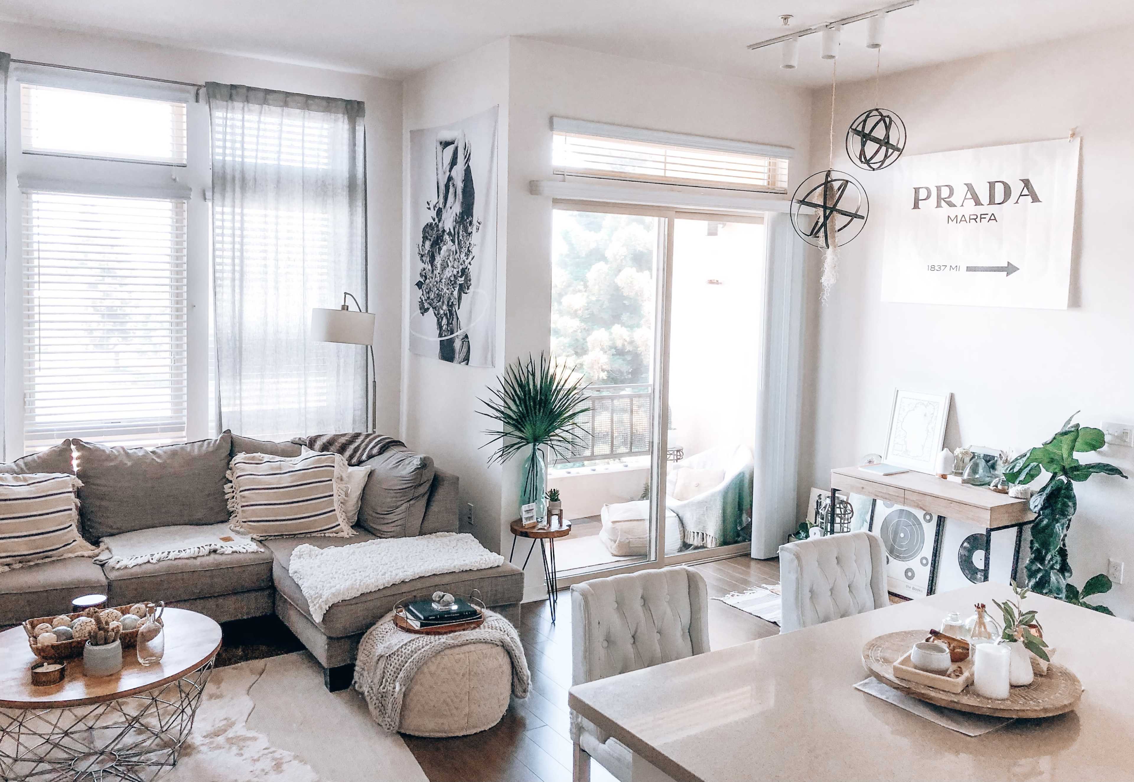 A modern living room with a large sectional sofa, a circular coffee table, and a dining area featuring a light-colored table and upholstered chairs, with a view of a balcony through sliding glass doors.
