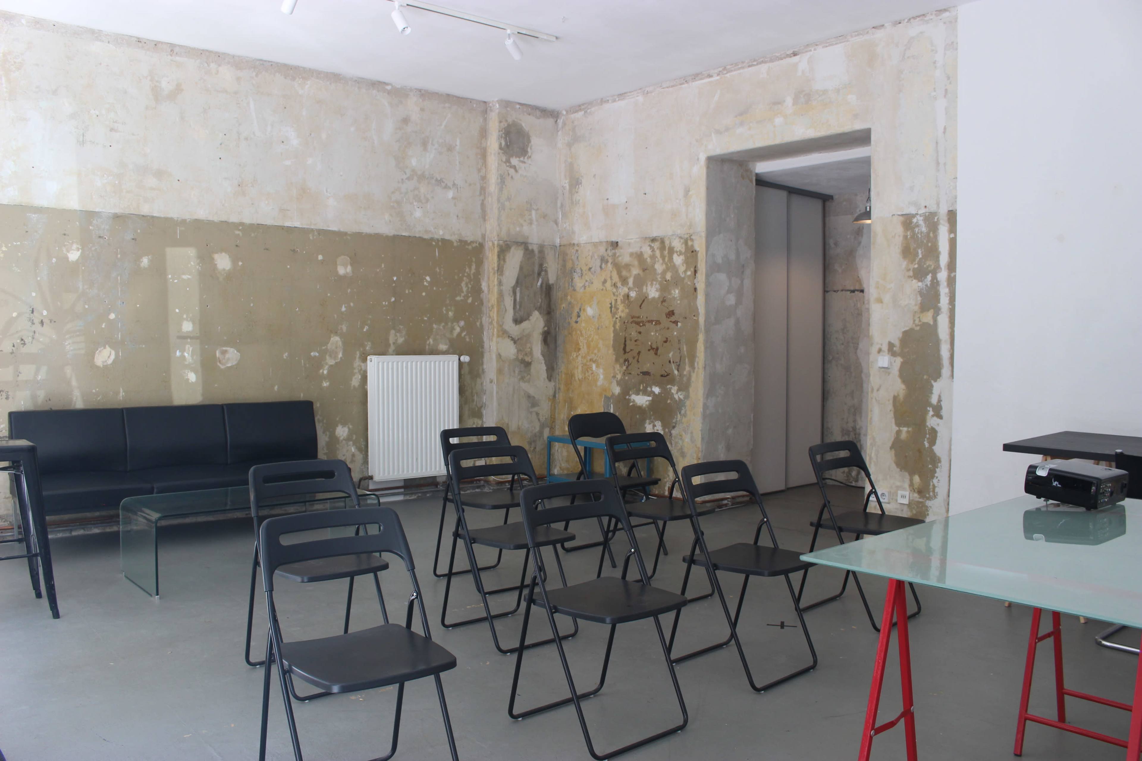 The image shows a sparse room with an unfinished wall, a black sofa, several black folding chairs arranged in rows, and a glass table on a red base.