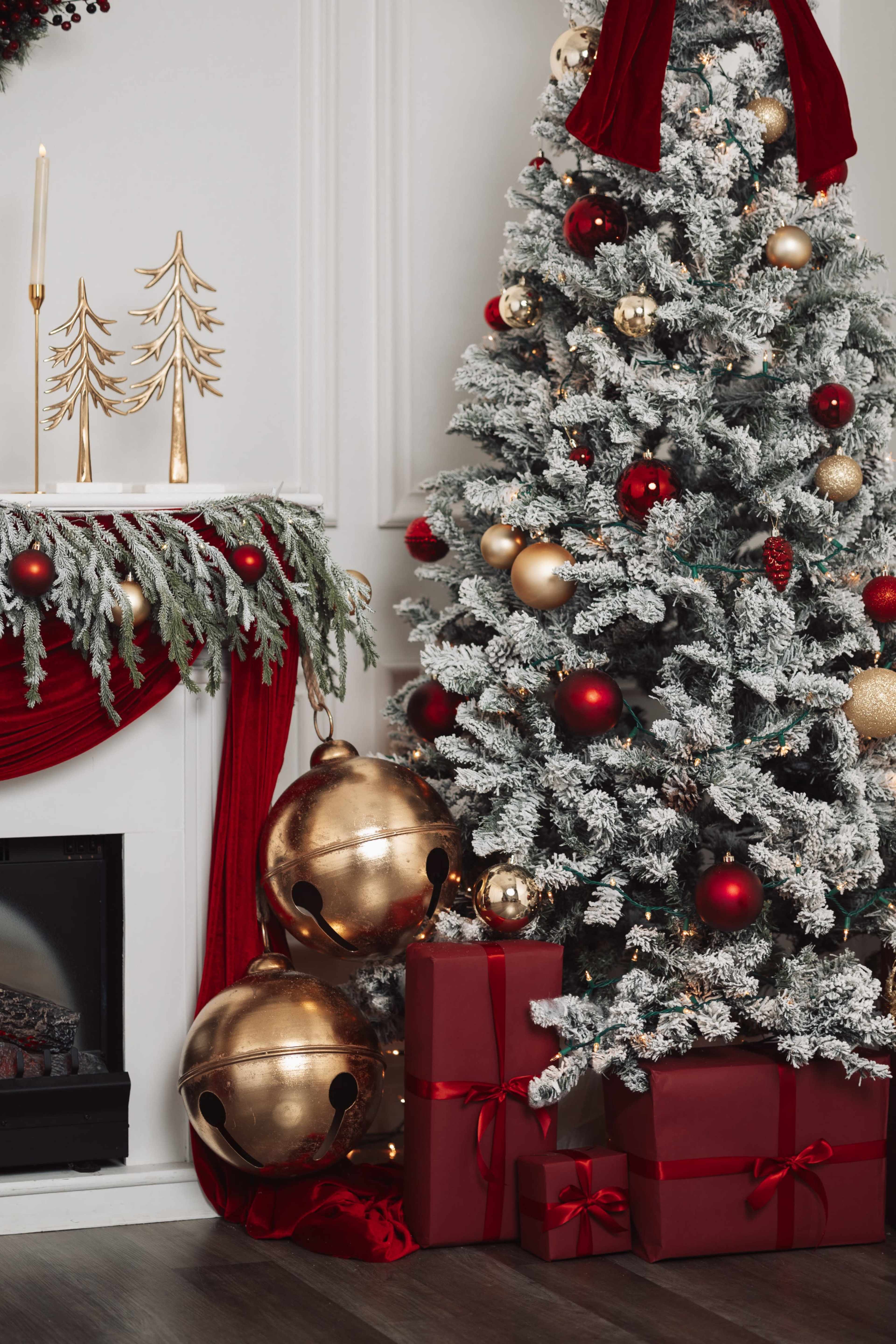 A decorated Christmas tree with red and gold ornaments is placed beside a fireplace adorned with garlands and wrapped gifts.