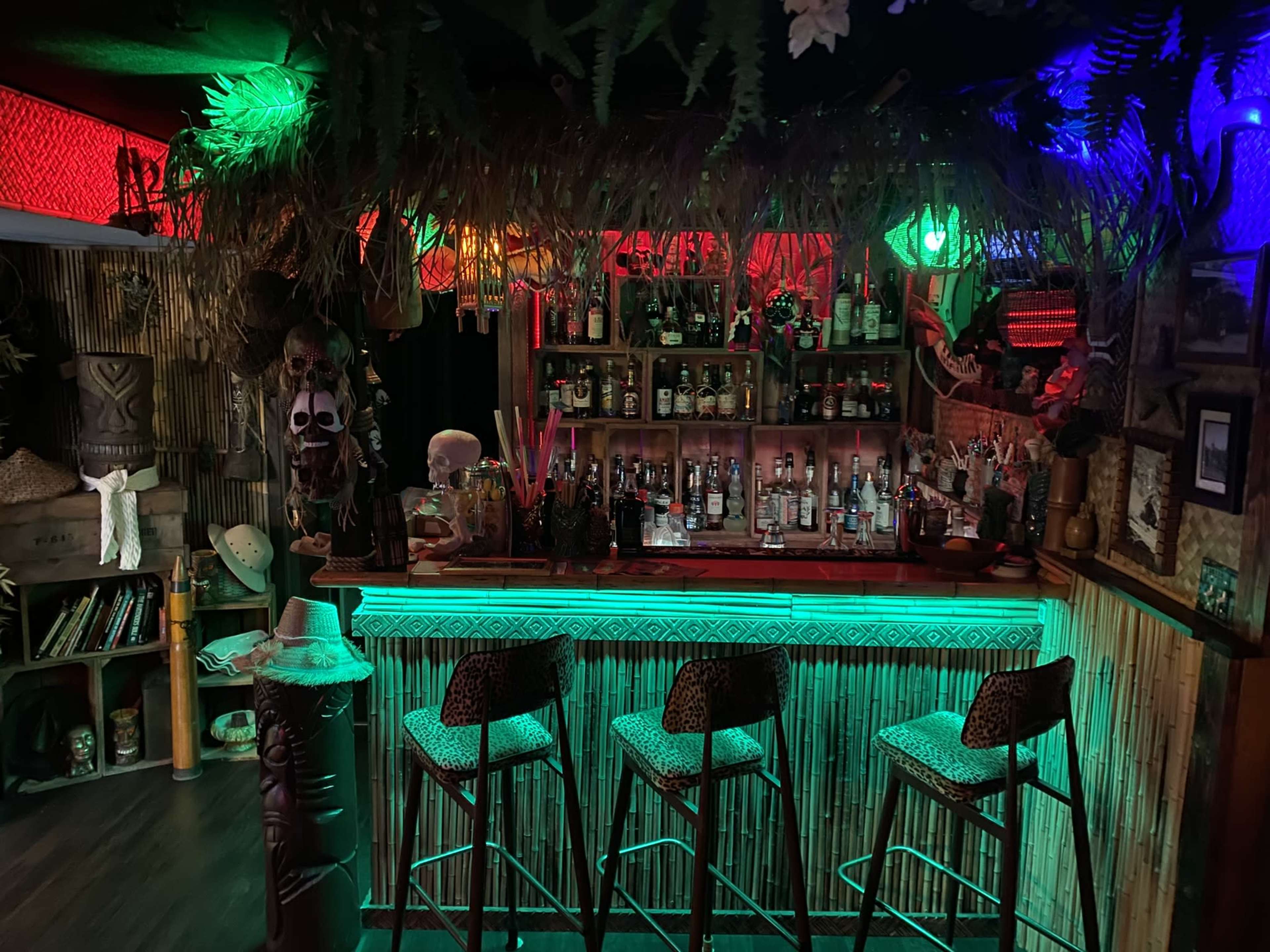 A tiki bar with a thatched roof, colorful lighting, and a variety of bottles displayed on the shelves behind the bar.