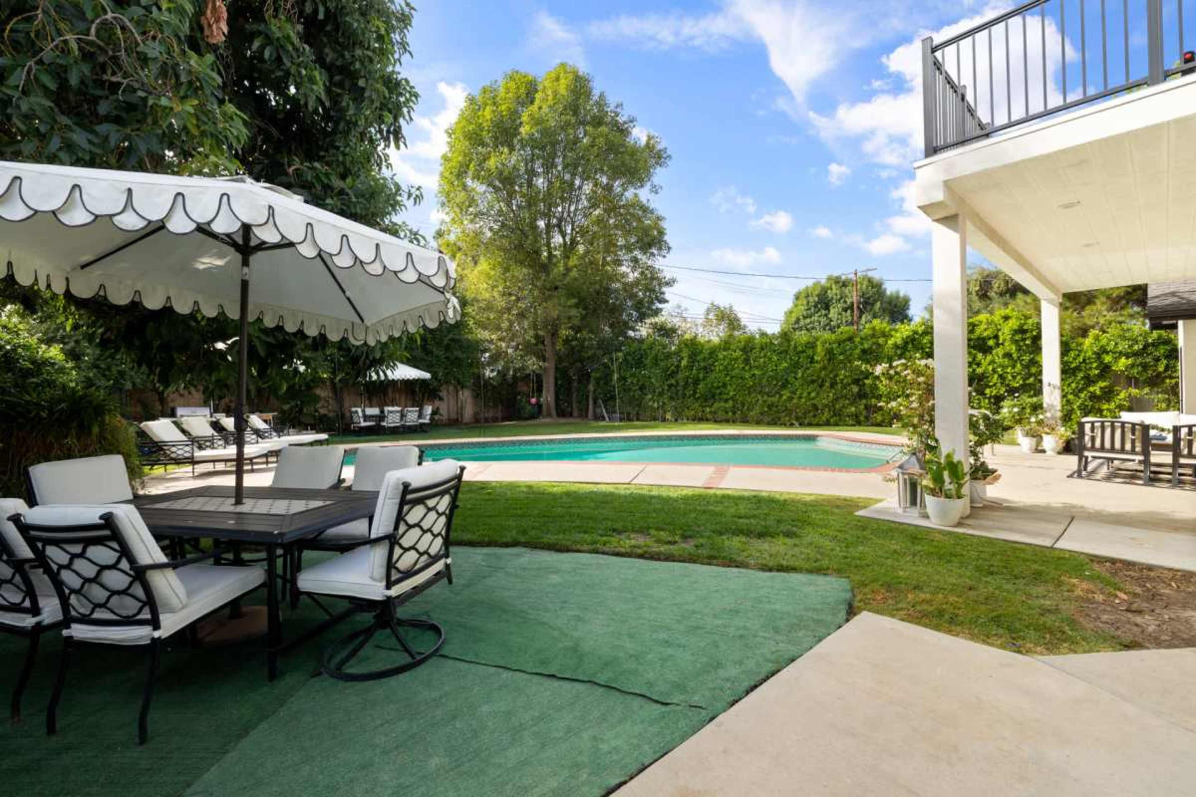 The image shows a backyard with a swimming pool, a dining table under an umbrella, and green landscaping.