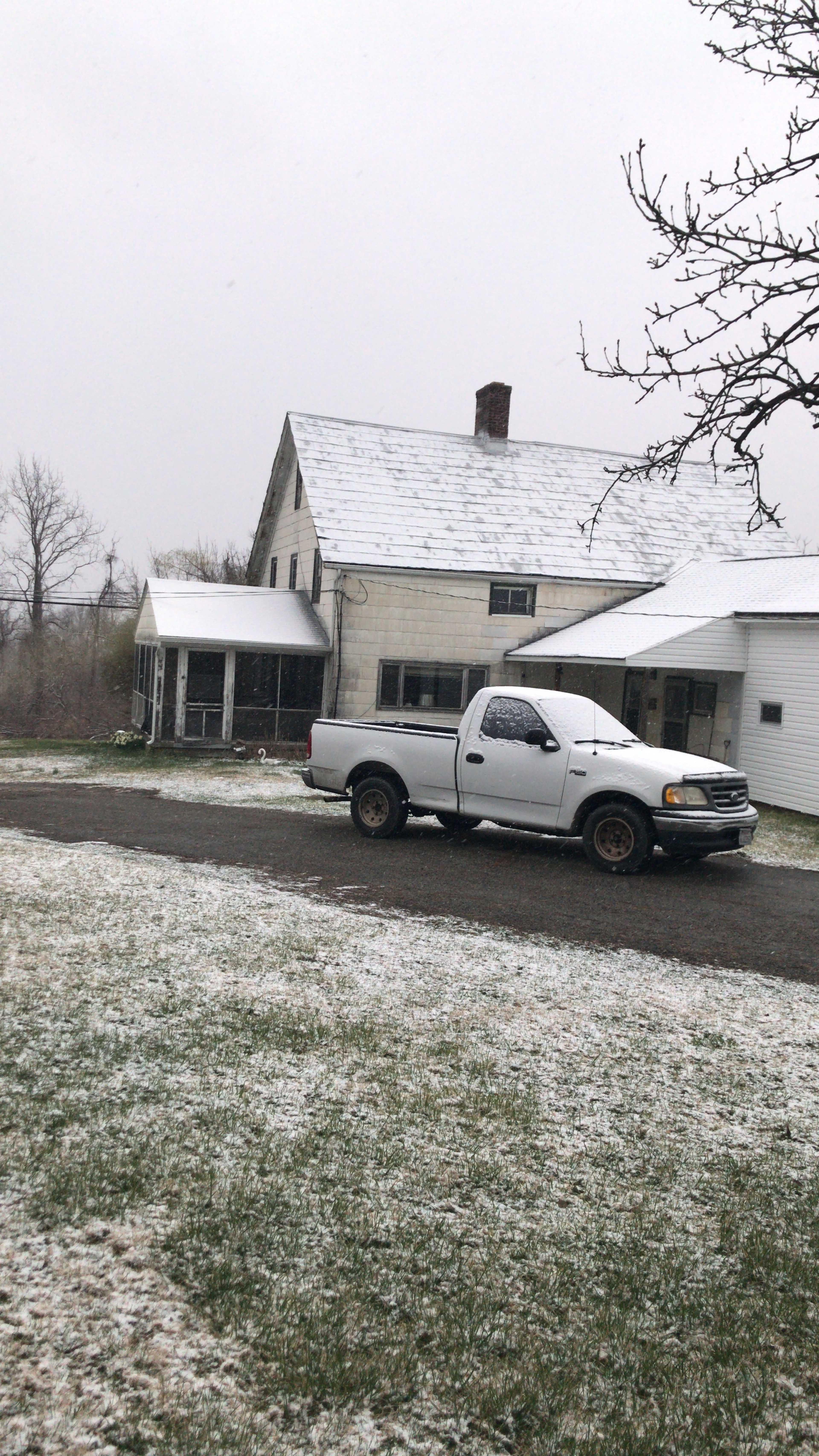 A white pickup truck is parked on a gravel driveway in front of a two-story house with a snow-covered roof and a light dusting of snow on the ground.
