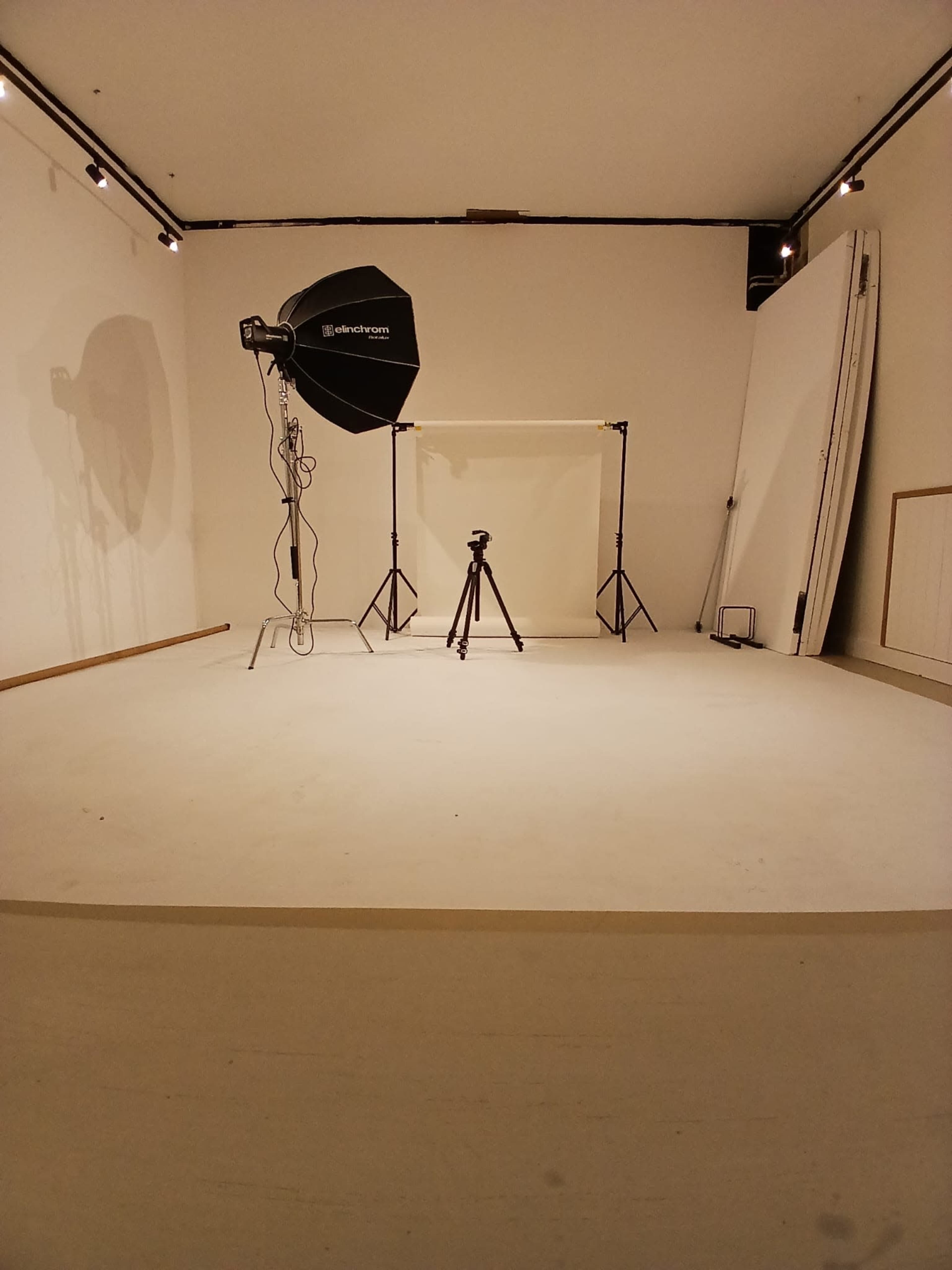 The image shows a photography studio with a tripod, softbox light, and a white backdrop set up in an empty room.