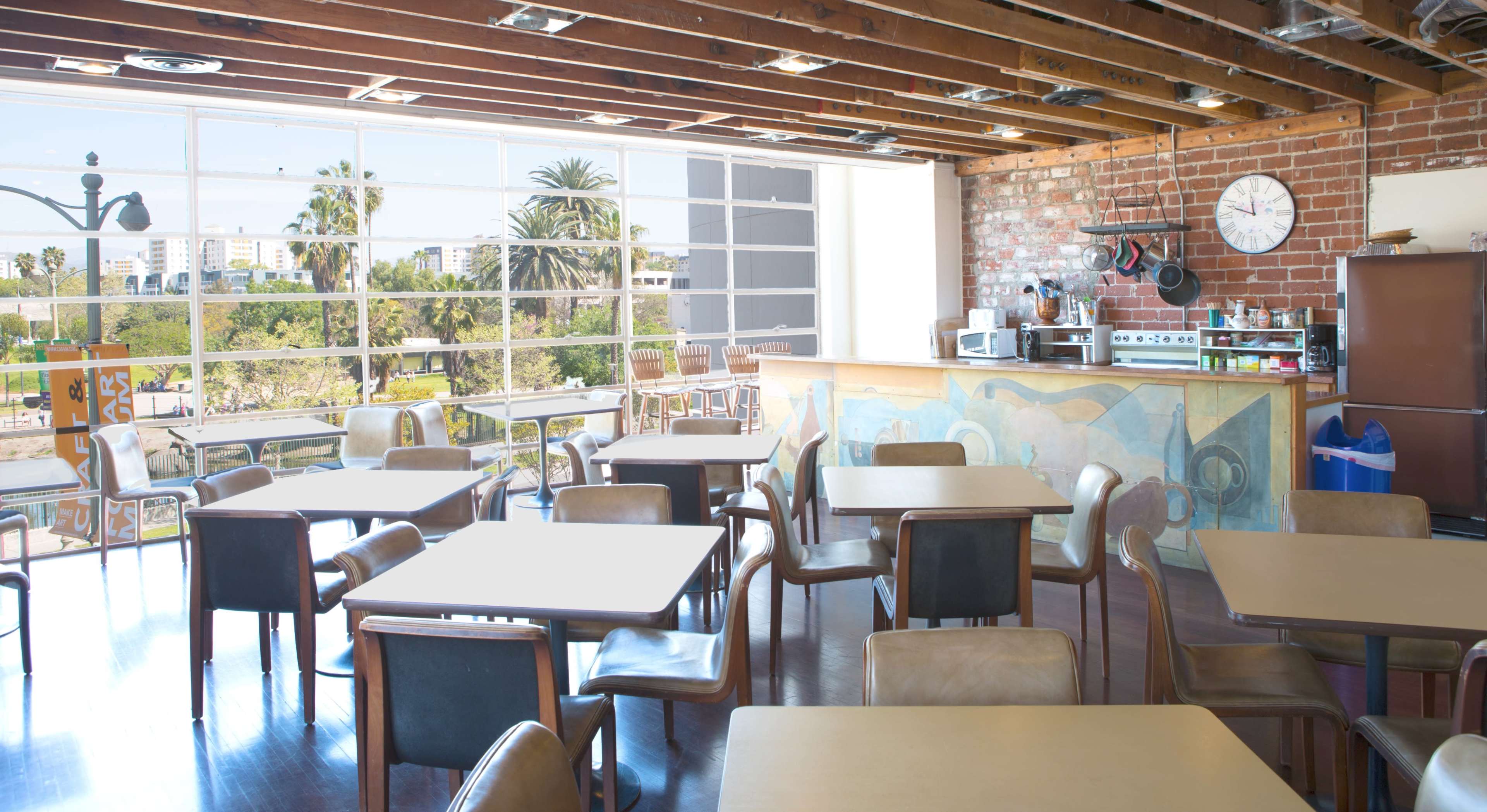 A bright, spacious café with wooden beams and large windows overlooking greenery, featuring several empty tables and a counter with kitchen equipment.