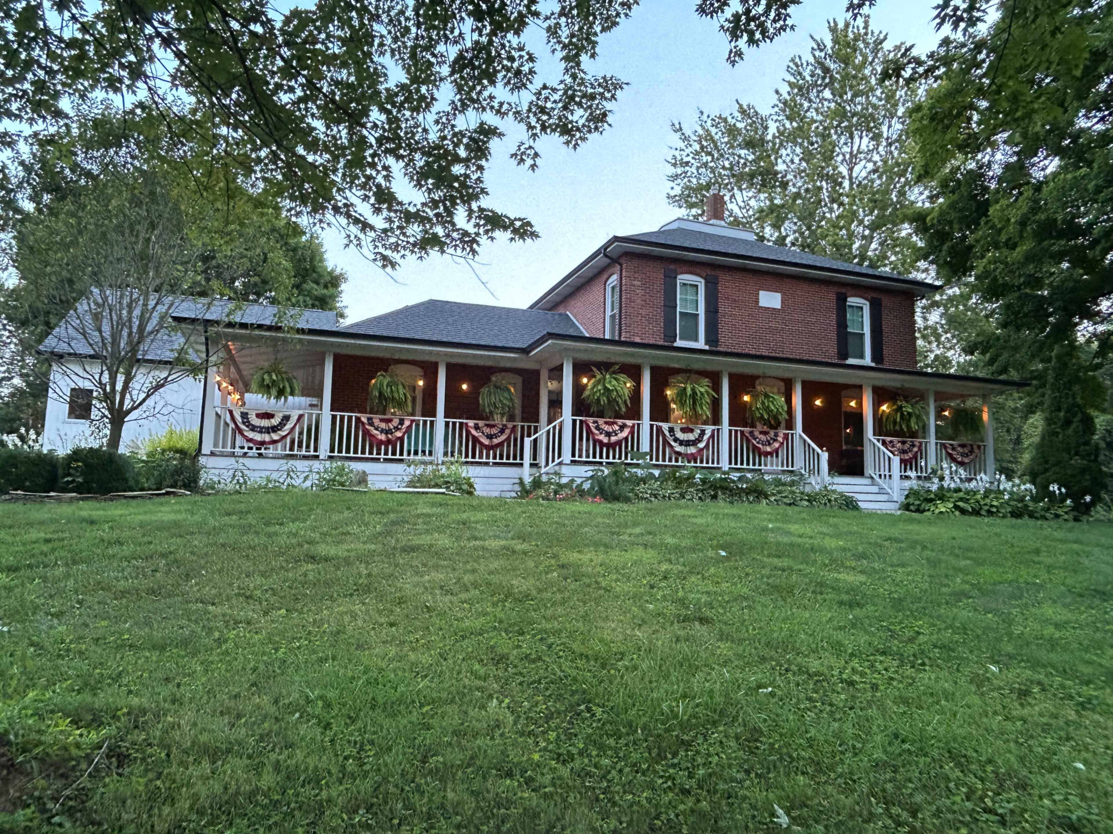 A two-story red brick house with a front porch adorned with hanging planters and patriotic bunting is framed by a lawn and trees.