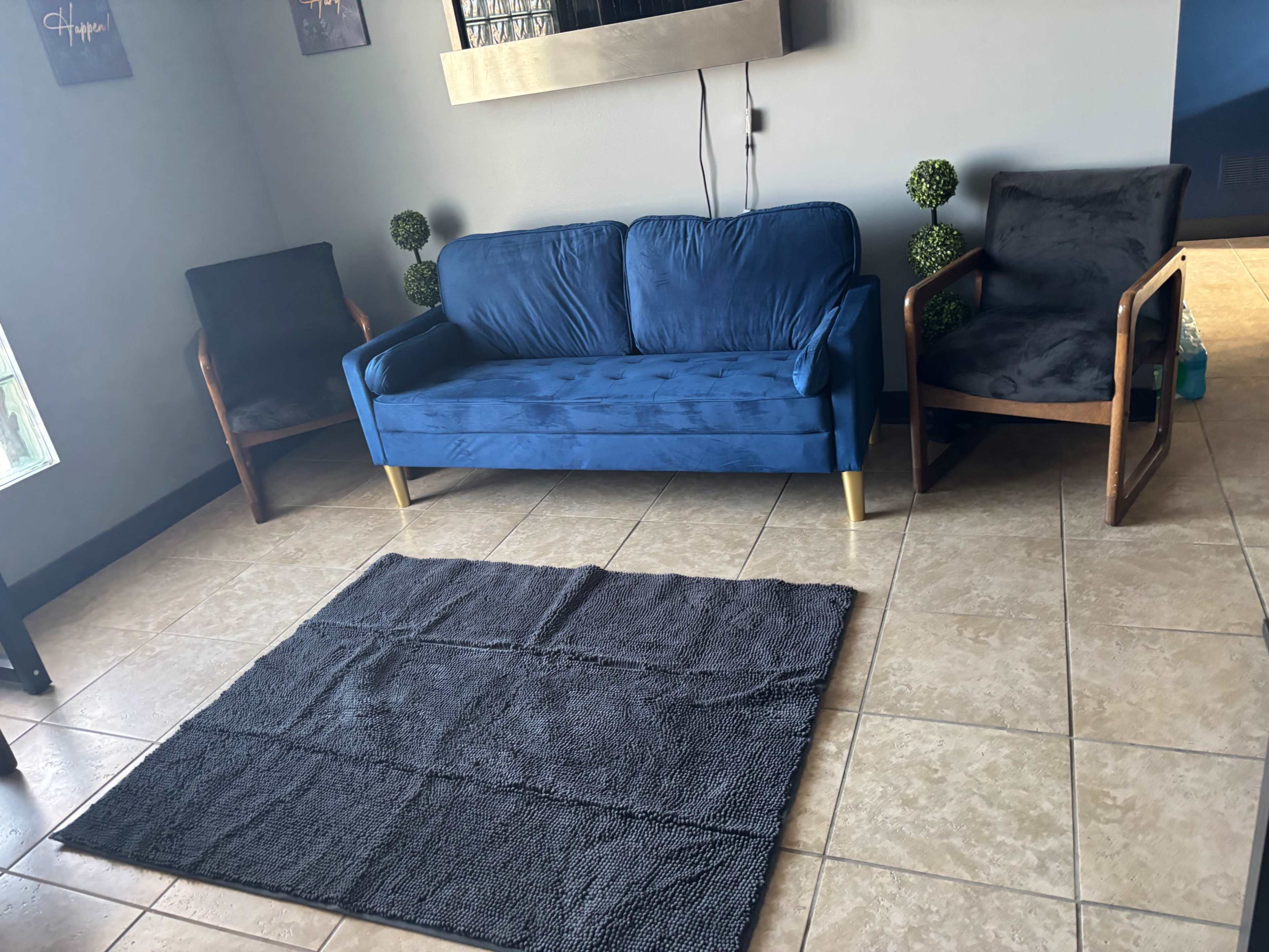 A blue sofa is positioned in the center of a room with two gray upholstered chairs and a textured black rug on the tiled floor.