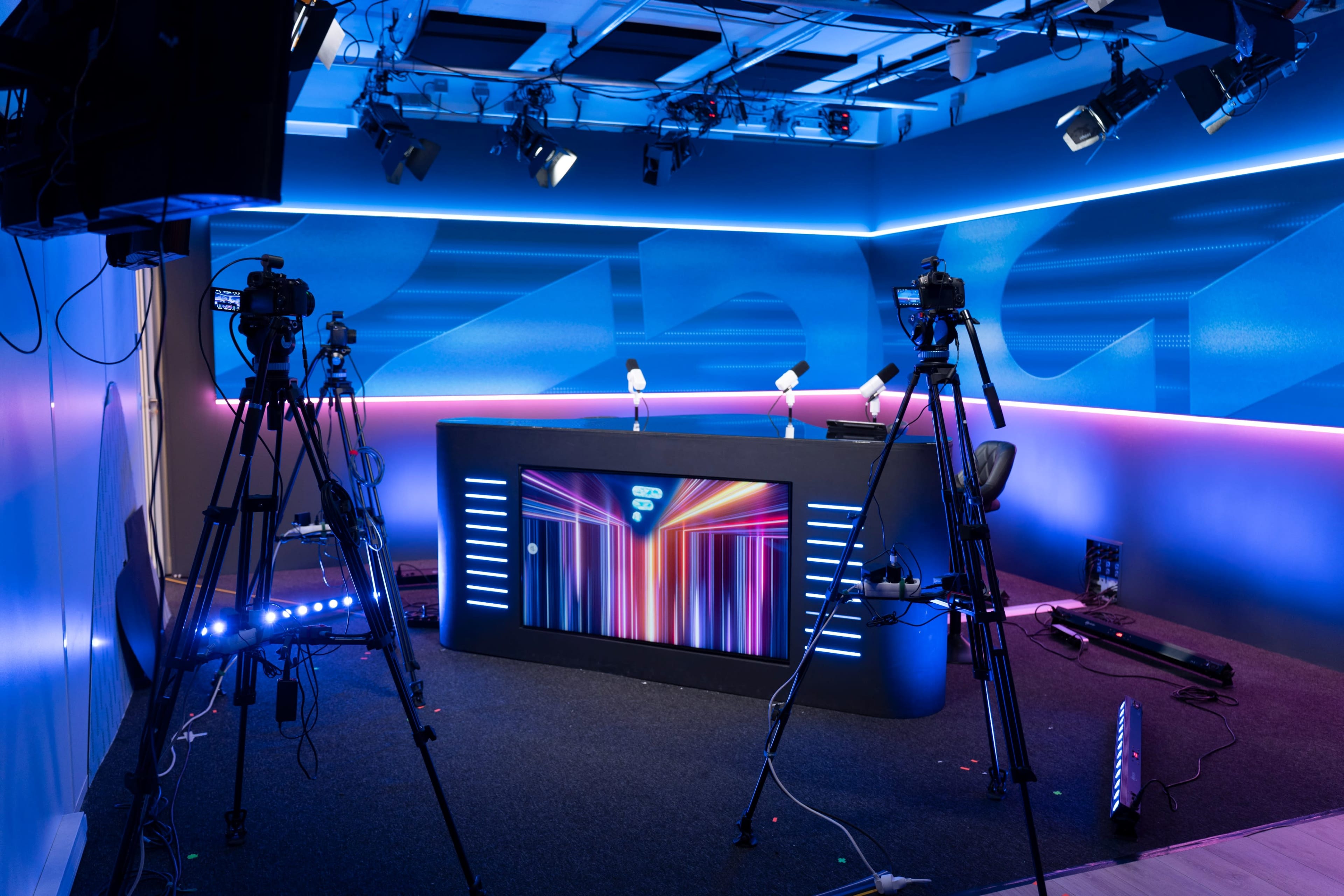 A brightly lit television studio features a central desk with colorful lighting and multiple cameras positioned around it.