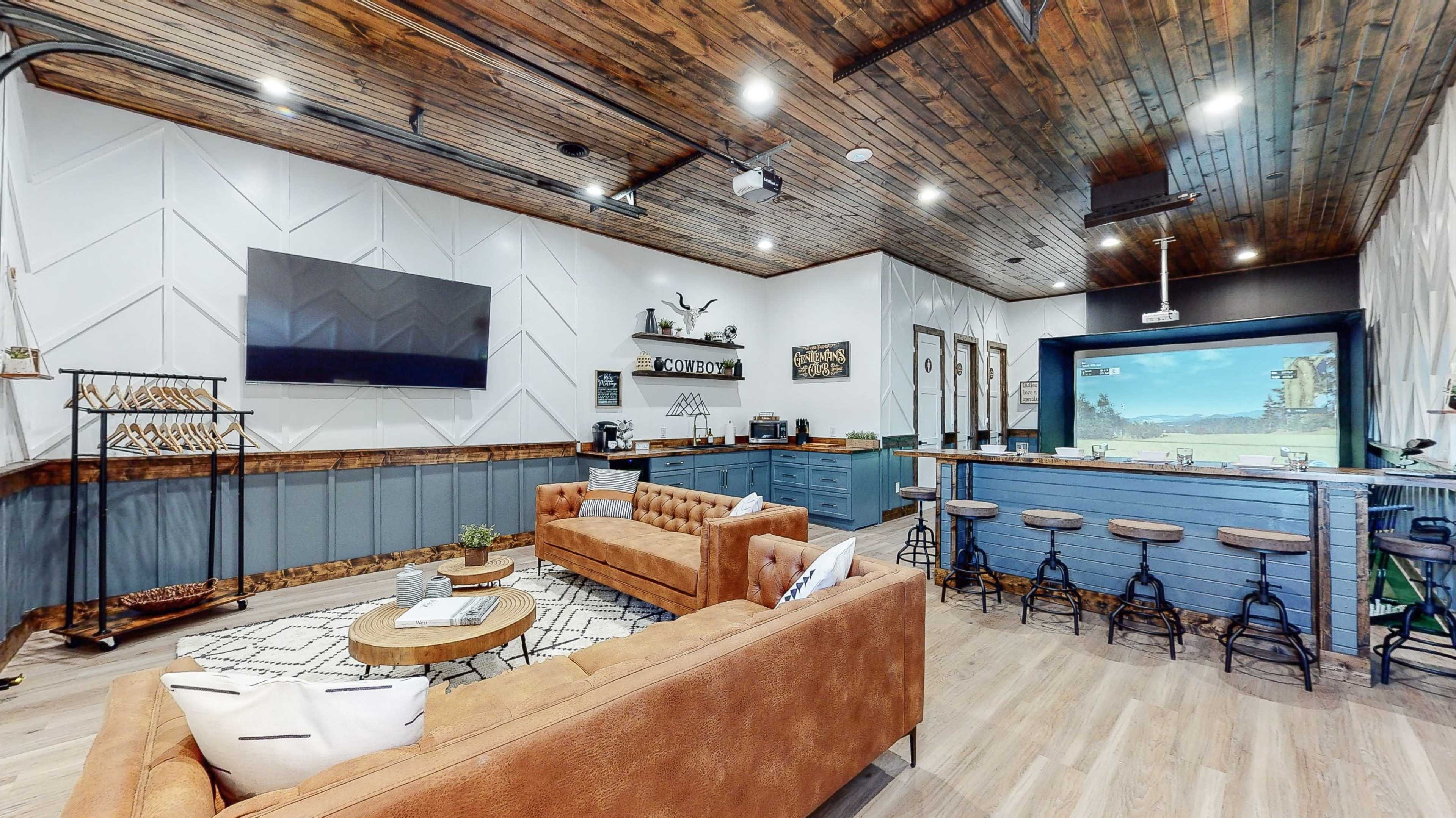 A modern living area with a brown tufted sofa, a large television mounted on the wall, a blue kitchenette in the background, and a rustic bar area with stools.