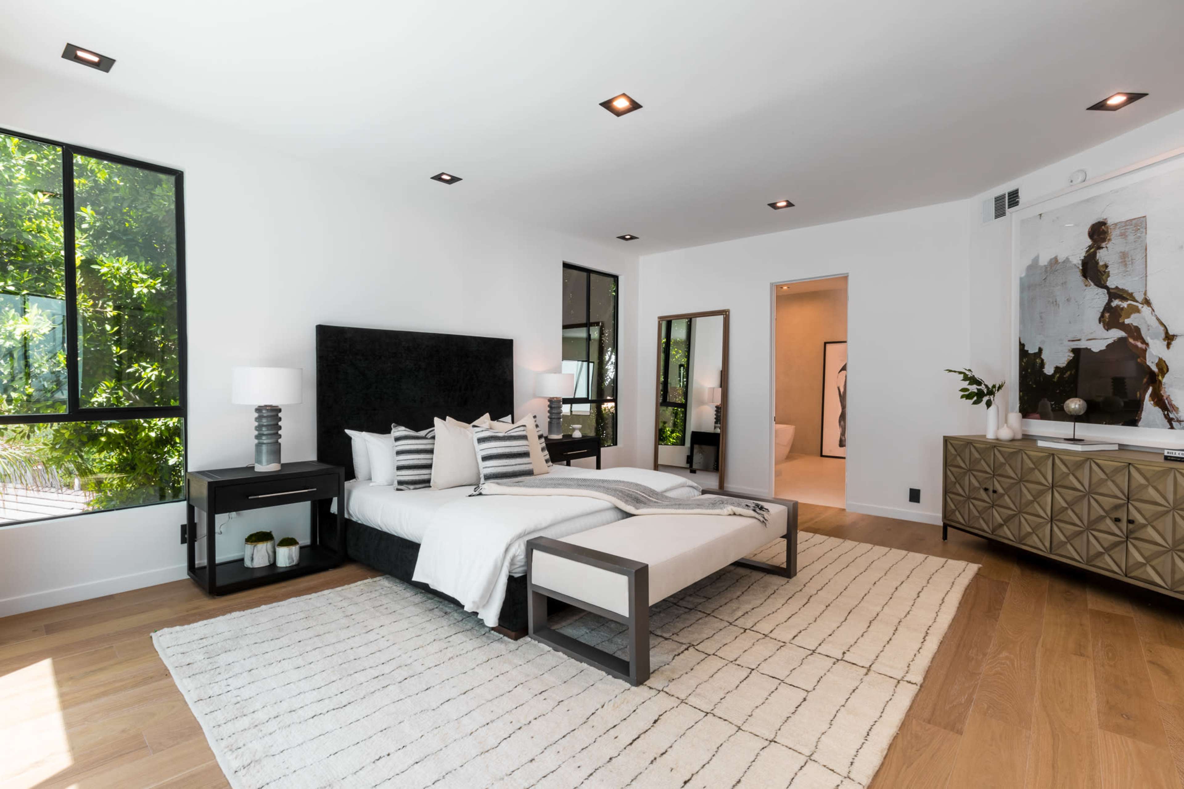 A modern bedroom featuring a black upholstered bed, two bedside lamps, a large mirror, and a neutral color palette with wooden flooring.