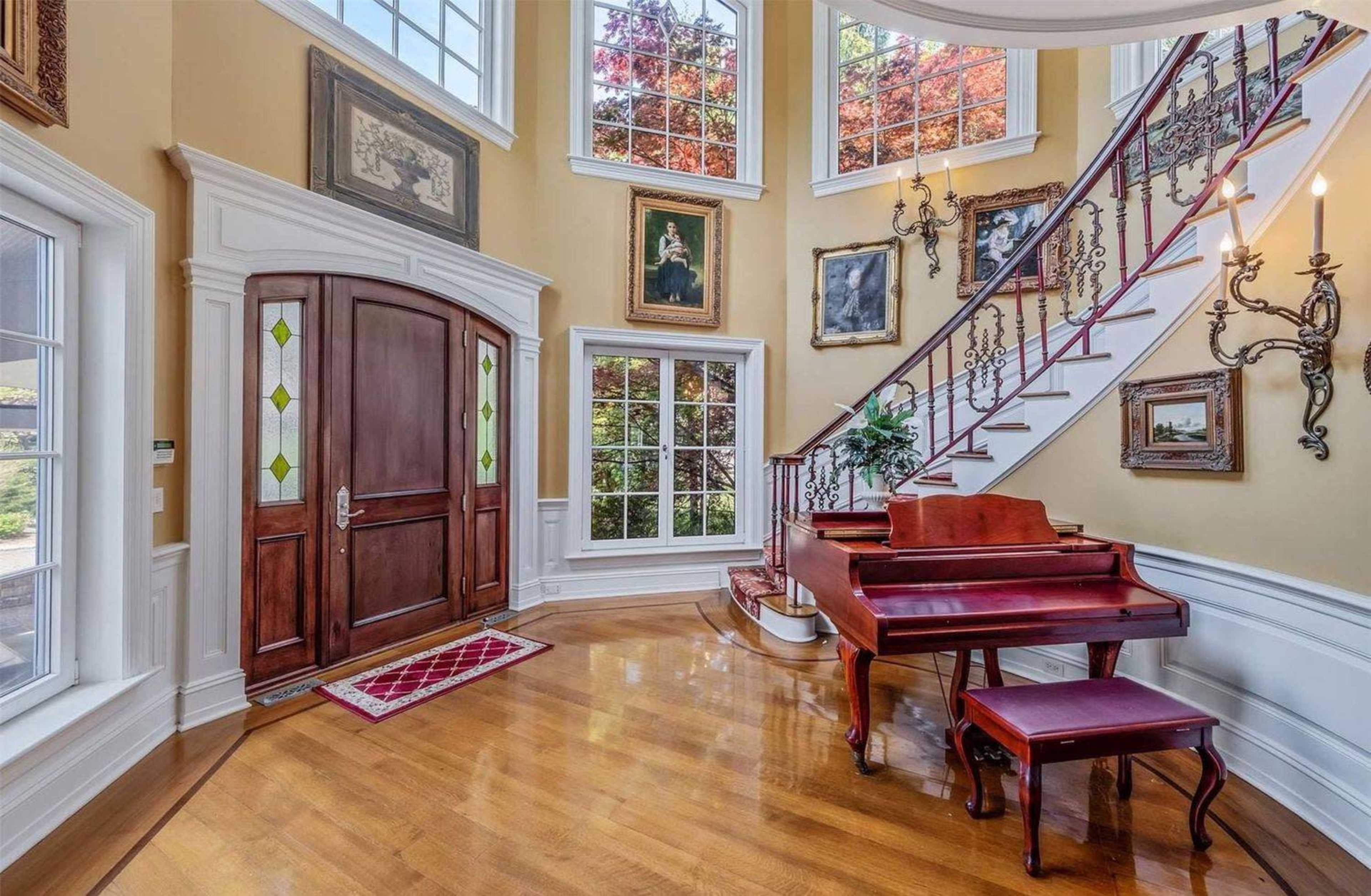 The image shows a spacious foyer featuring a wooden front door, a grand staircase, large windows, and a piano adjacent to the steps.