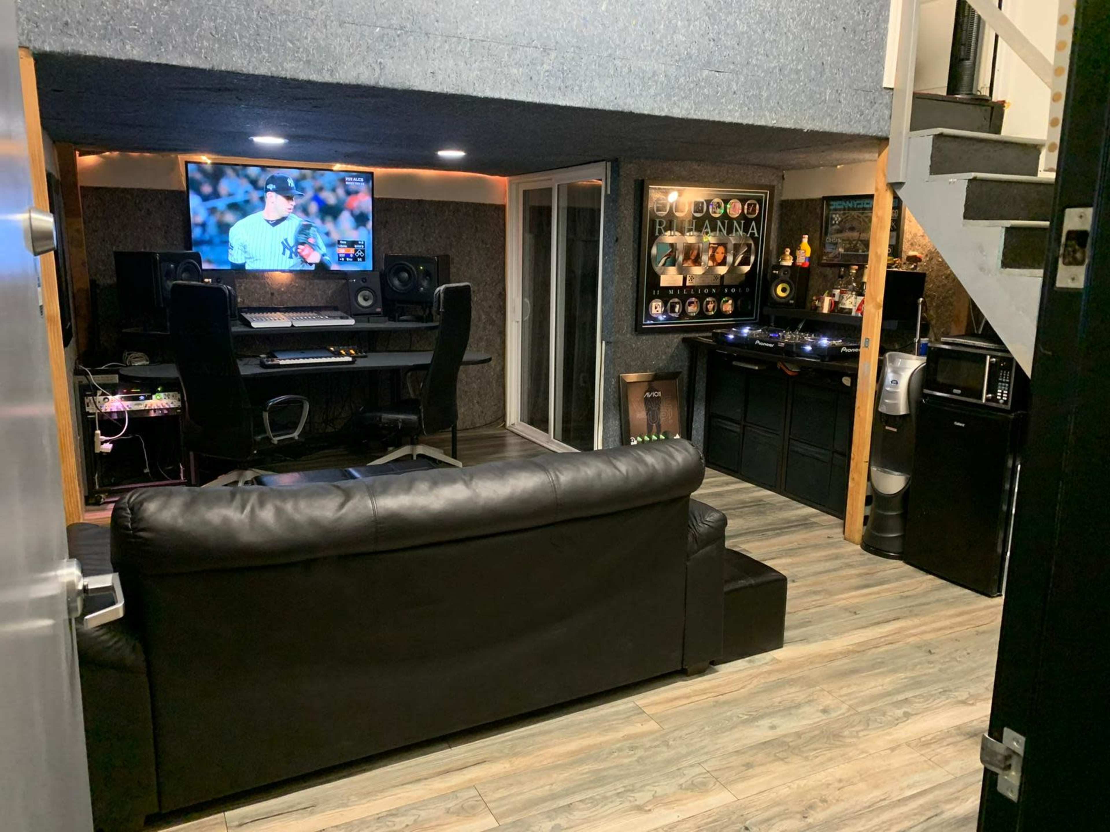 The image shows a modern basement studio with a large television, sound equipment, and a seating area featuring a black leather couch.