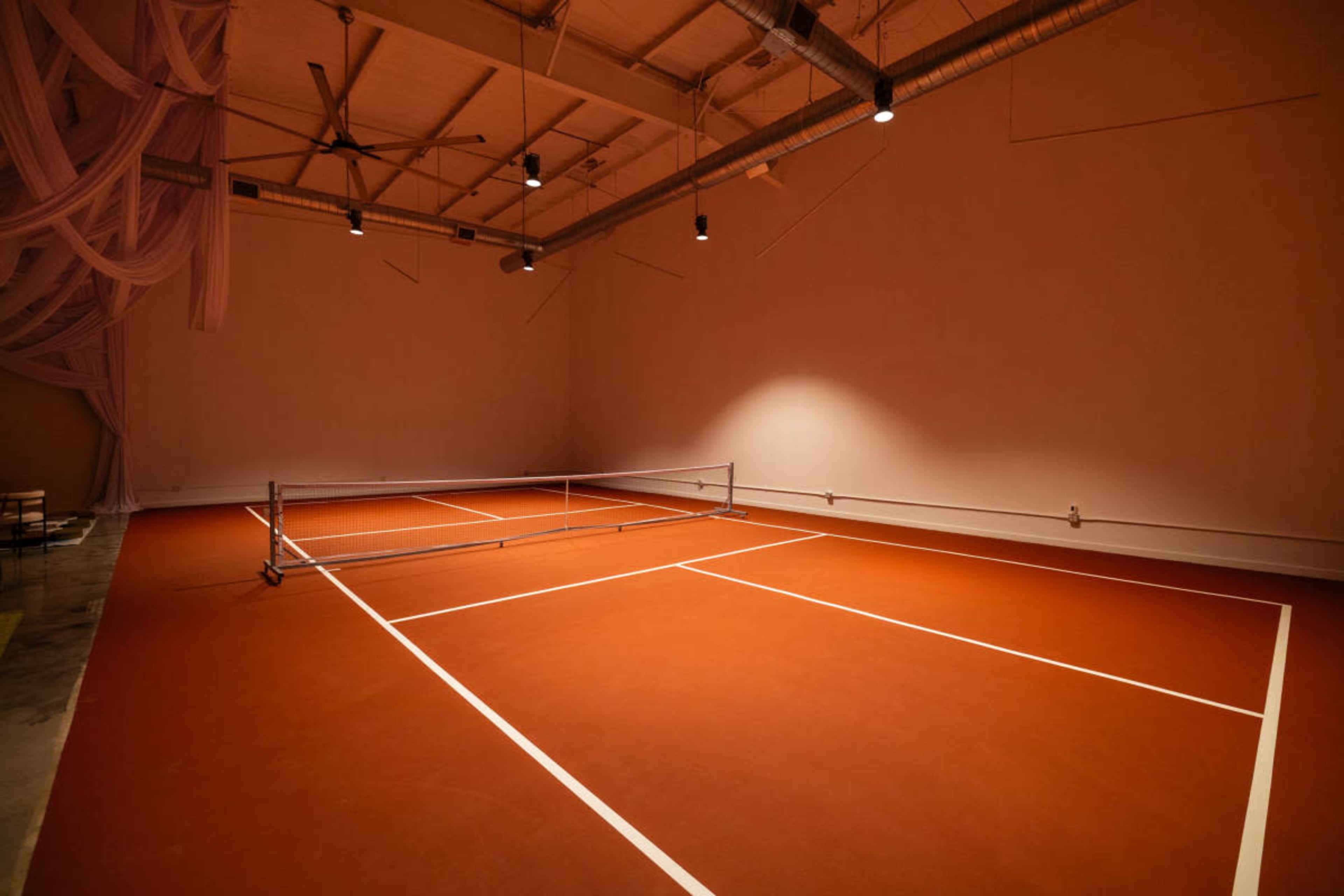 The image shows a sparsely decorated indoor space with a tennis court featuring a red surface and a net in the center.