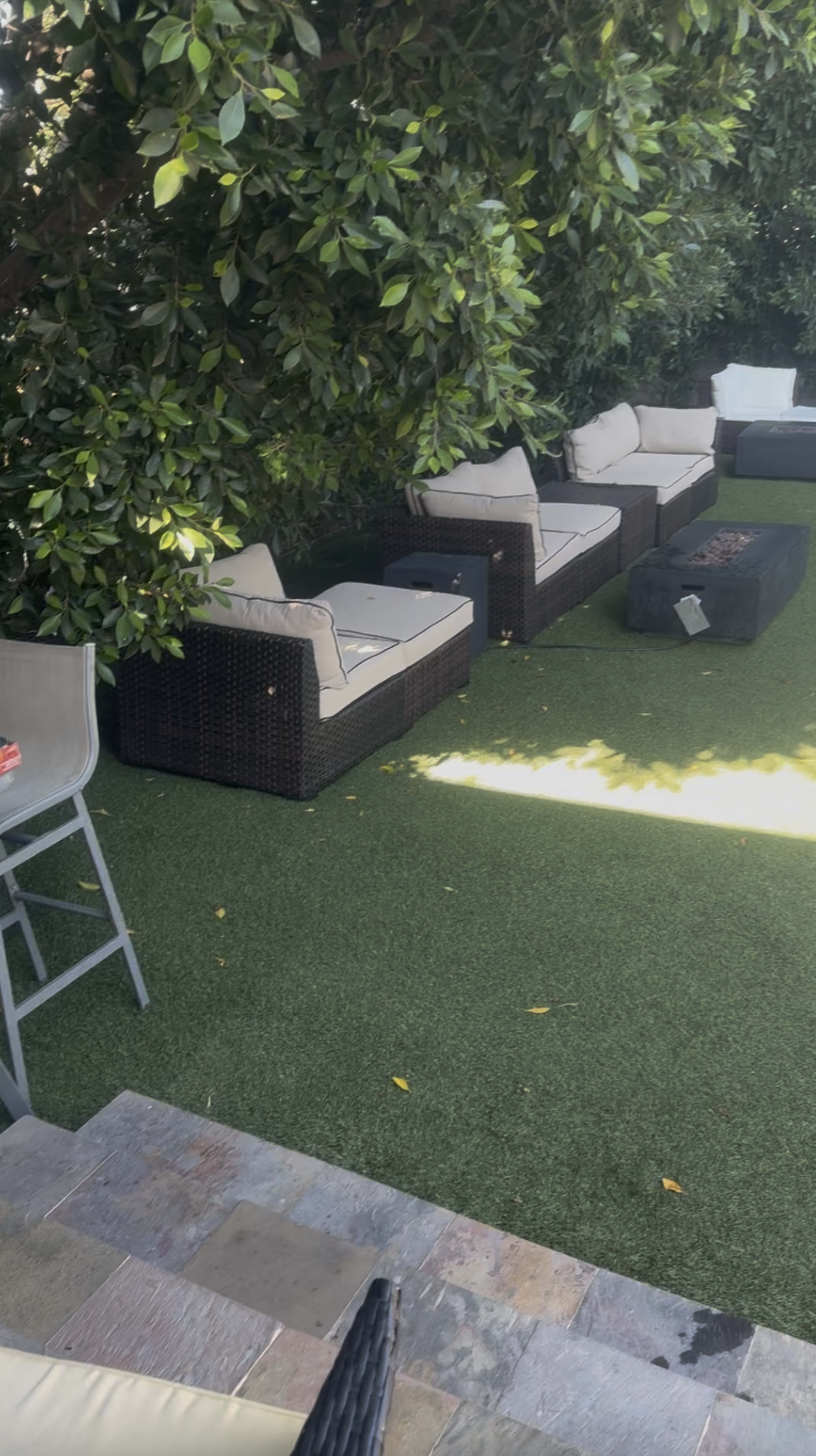 The image shows a small outdoor area with several patio chairs and a table arranged on artificial grass, surrounded by lush greenery.