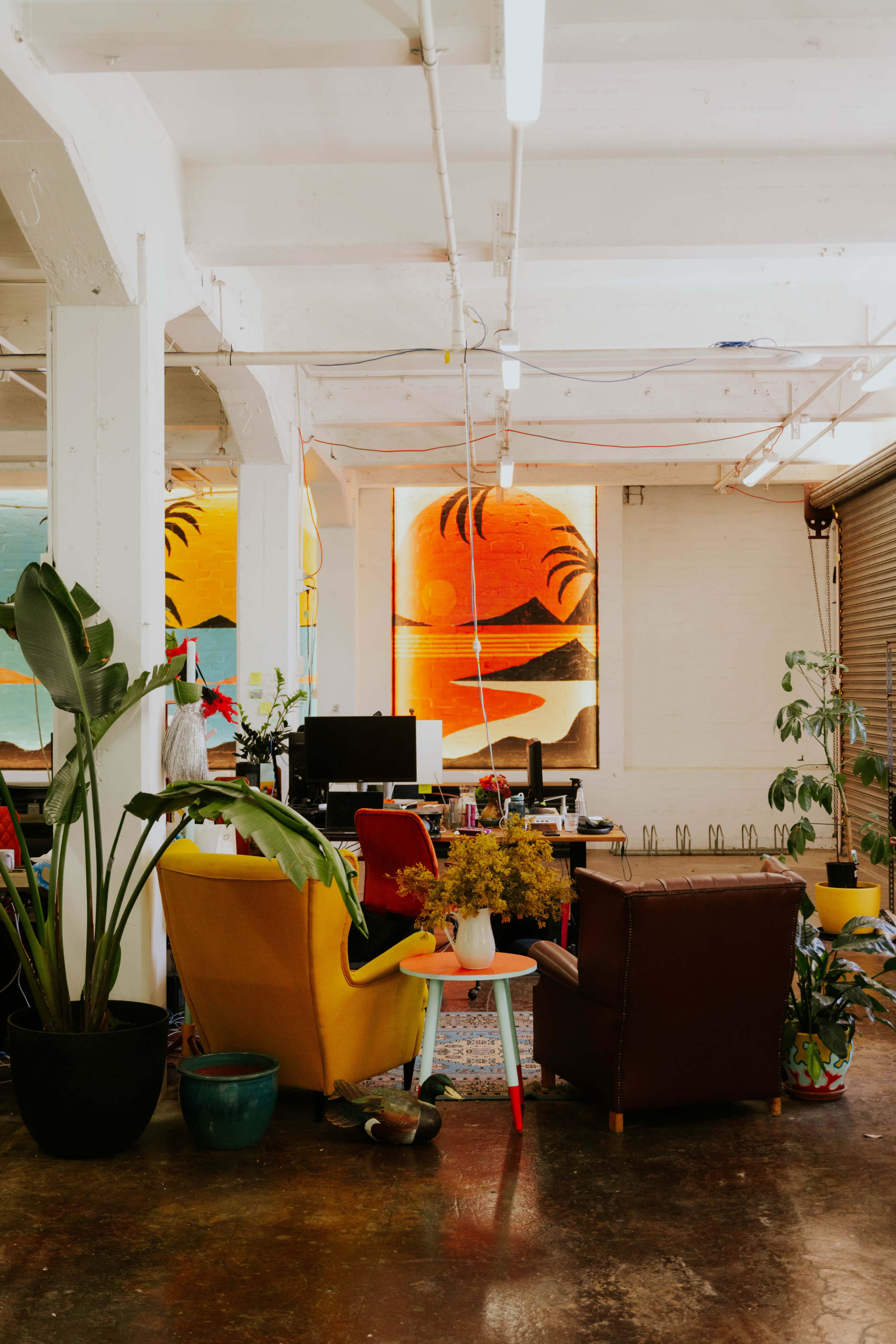 The image shows a stylish workspace with colorful chairs, plants, and large abstract artwork on the walls.