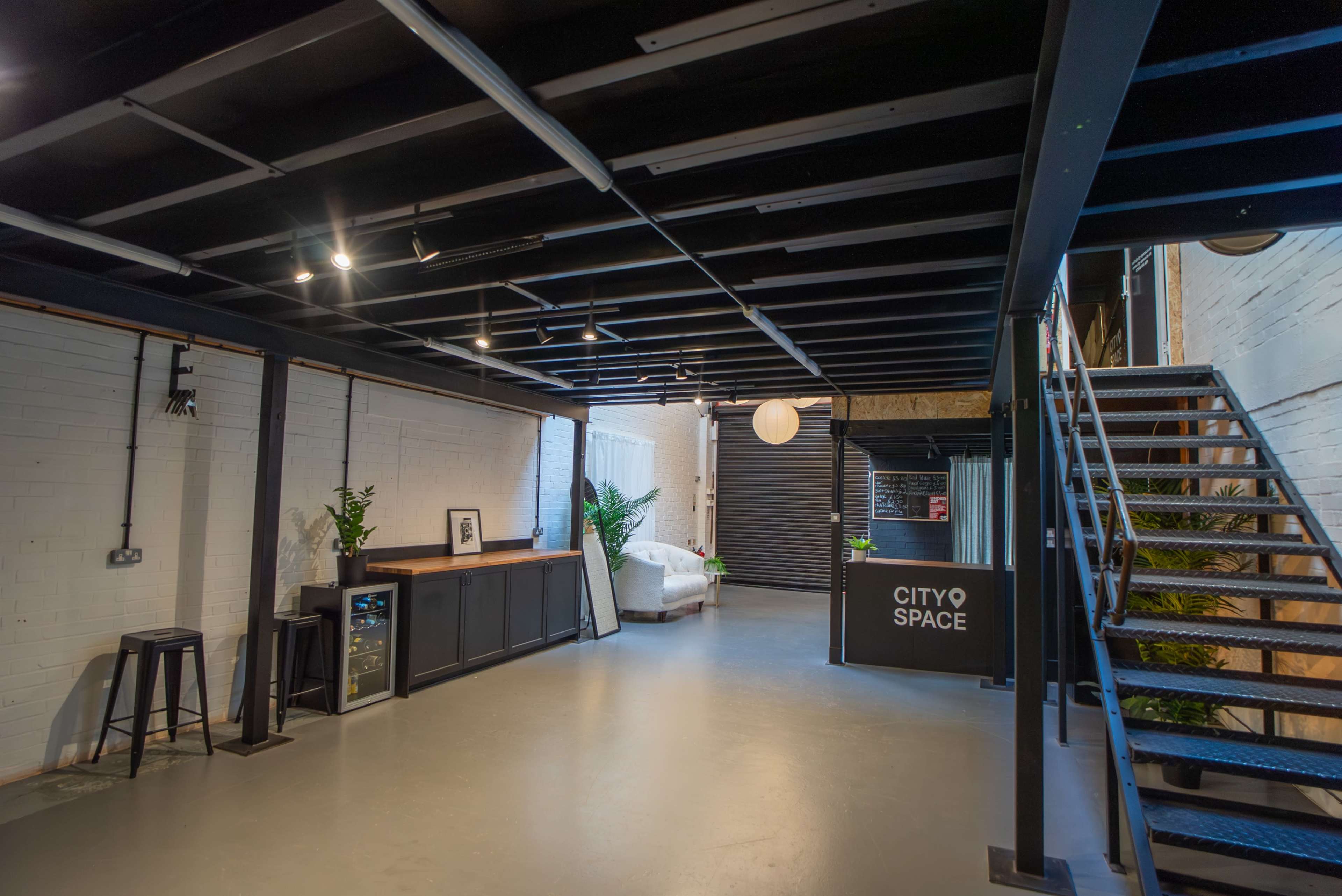 The image shows a modern, open interior space featuring a bar area, a staircase, and minimal furniture under a black beam ceiling.
