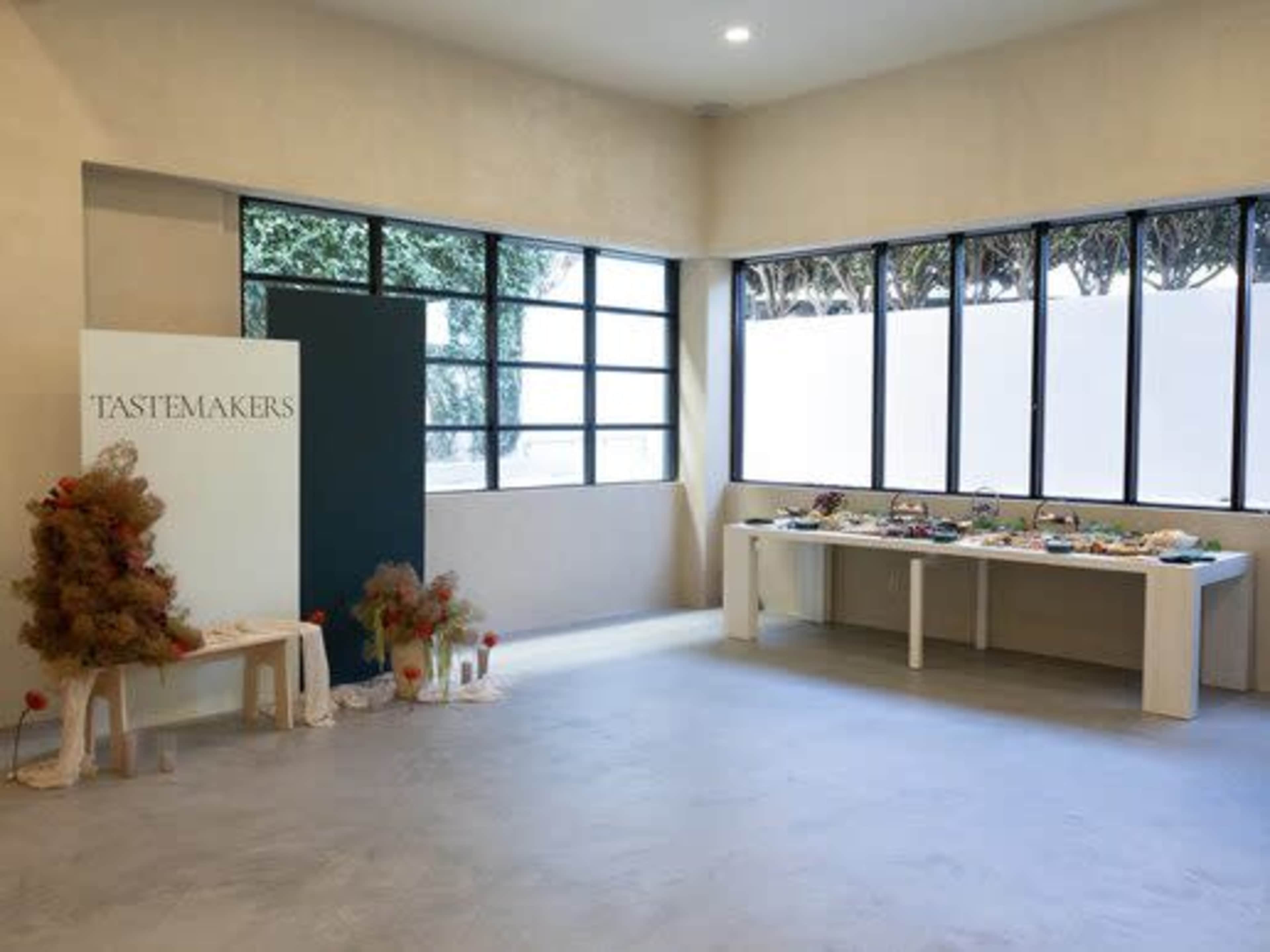 The image shows a spacious interior with large windows, featuring a display table set with various decorative items and a sign that reads "TASTEMAKERS."