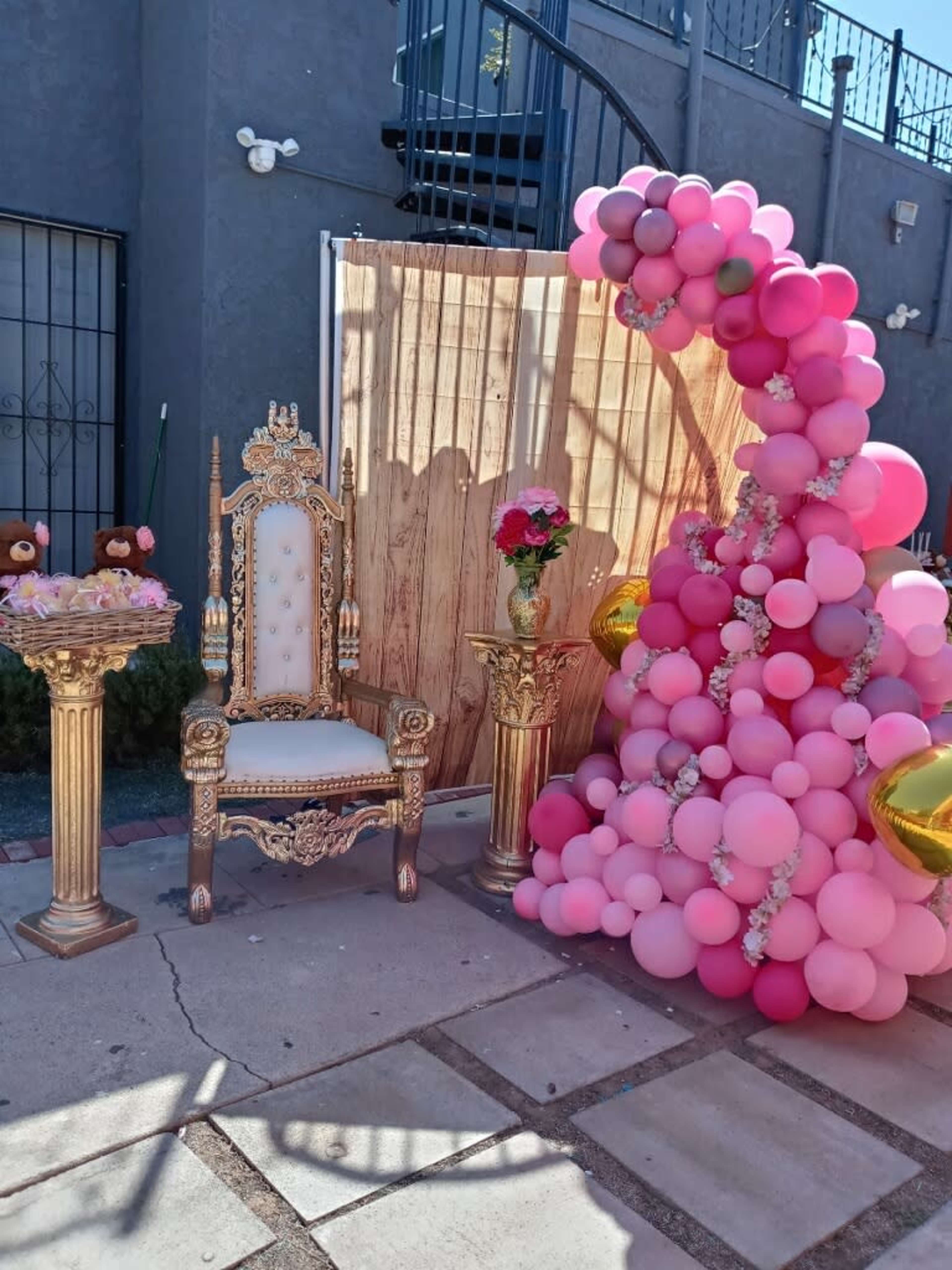A decorated party corner featuring a golden throne, two pedestal tables with floral arrangements, and a large arch of pink balloons.