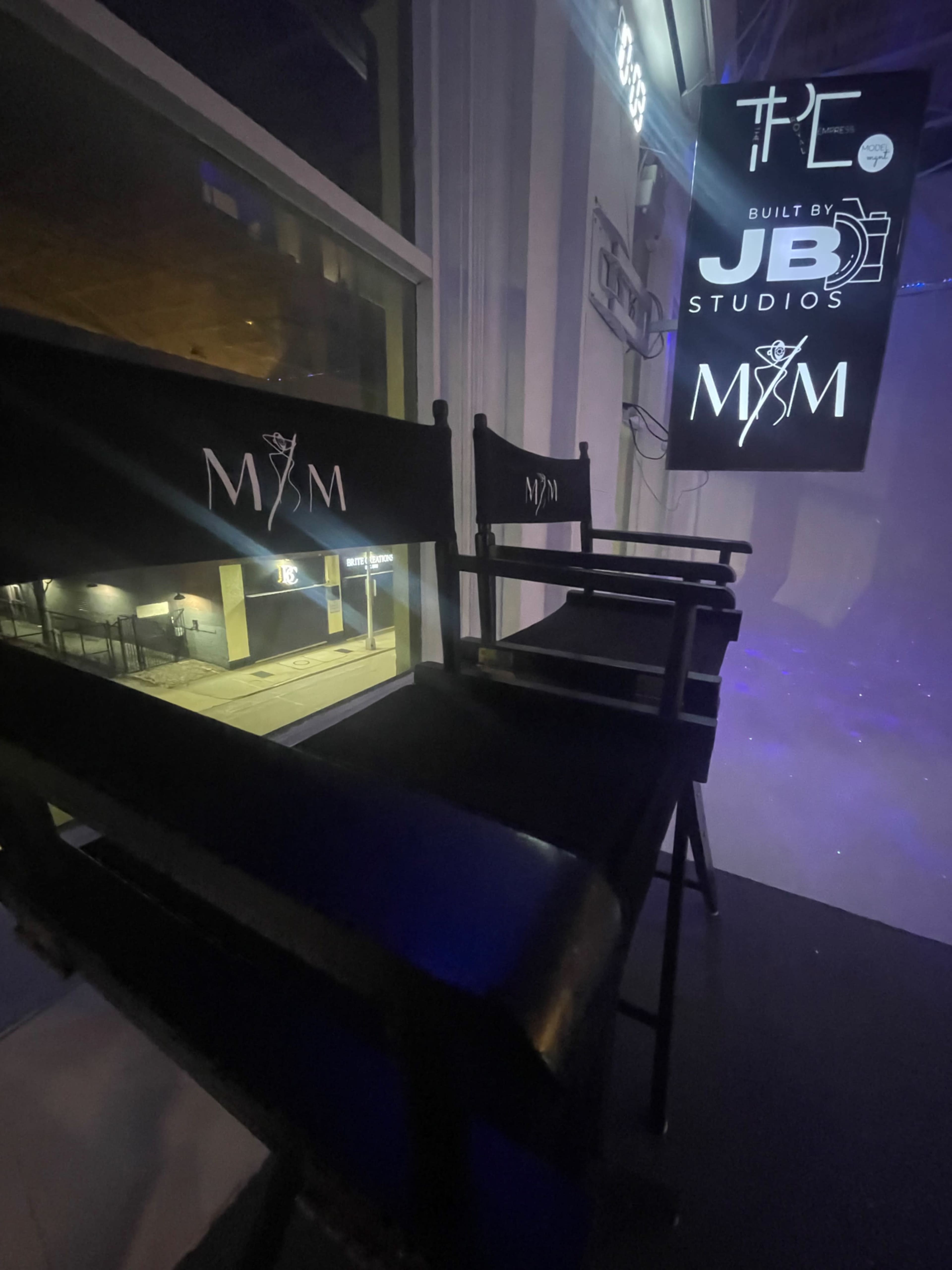The image shows two black director's chairs with "M&M" embroidered on them, positioned in front of a large illuminated sign that reads "THE M&M" and "BUILT BY JB STUDIOS."