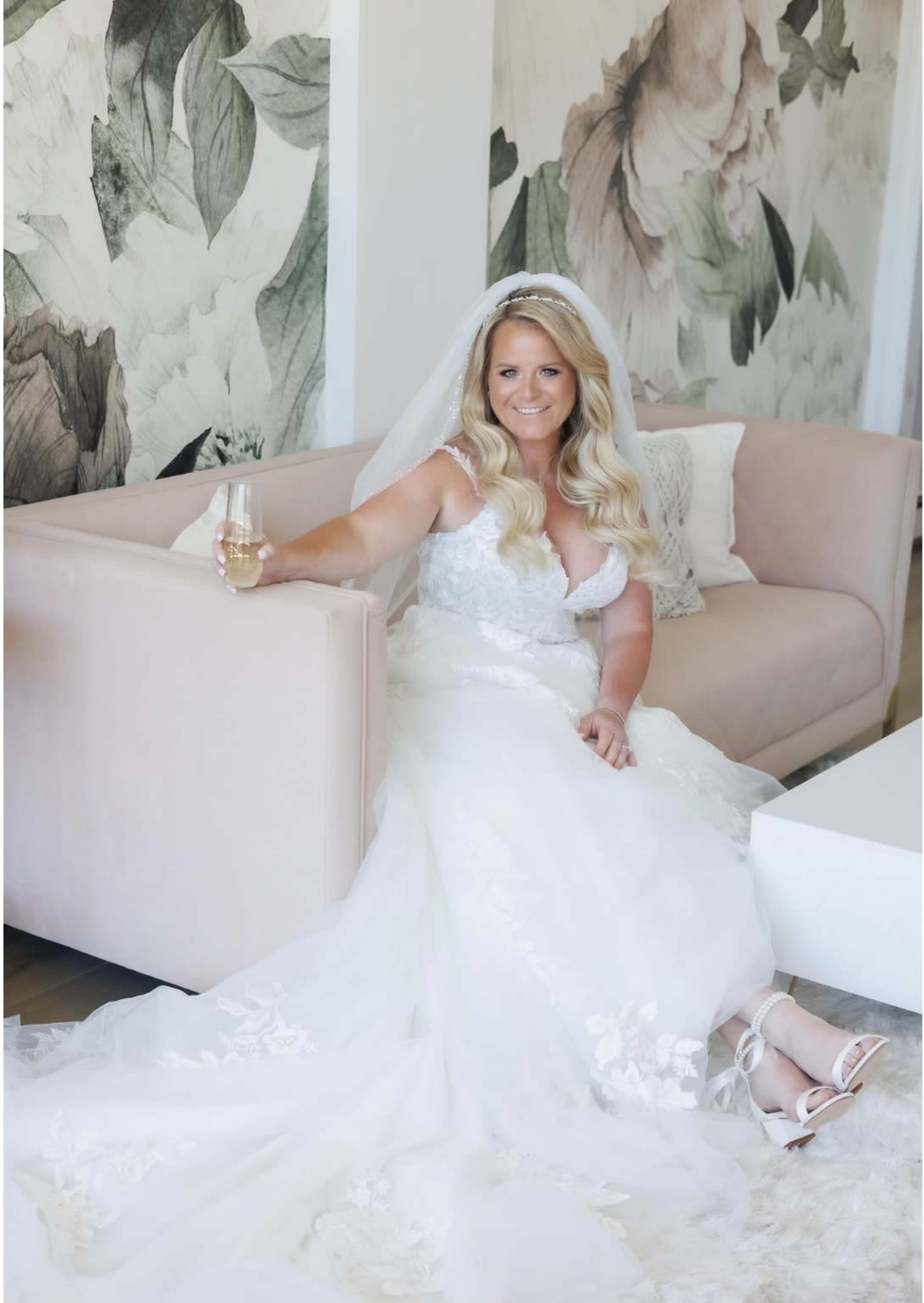 A bride in a white wedding dress sits on a pink couch, holding a glass of champagne, with floral wallpaper in the background.